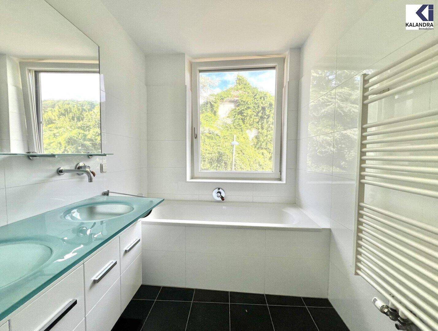 Modernes Badezimmer mit Badewanne, Doppelwaschbecken und Fenster mit Blick ins Grüne.