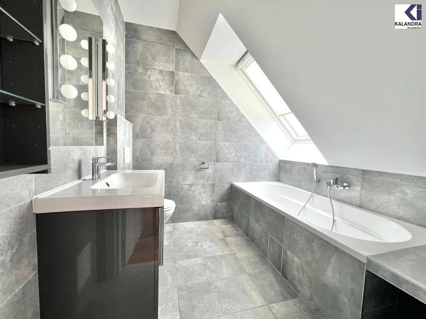 Modernes Badezimmer mit grauen Fliesen, Badewanne, Einzelwaschbecken und Dachfenster.