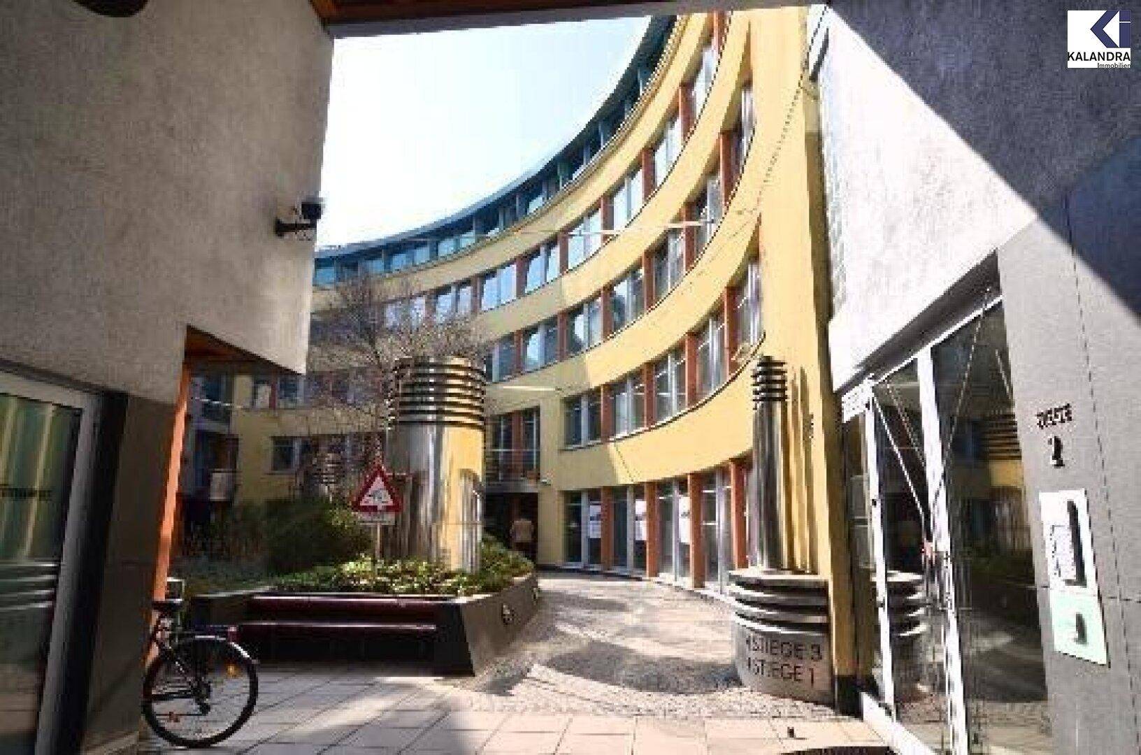 Geschwungene Fassade eines gelben Gebäudes mit vielen Fenstern, gesehen von einem Durchgang.