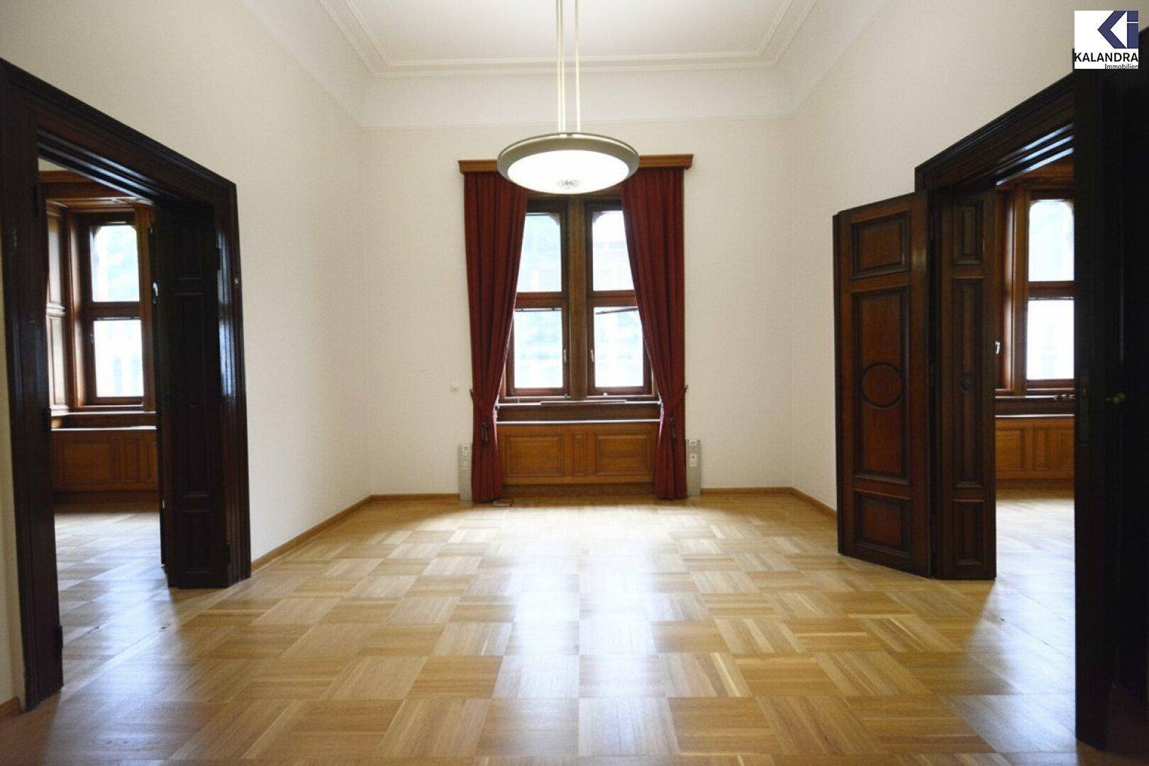 Großer, heller Raum mit klassischem Parkettboden, hohen Decken und eleganten Holztüren und Fenstern.