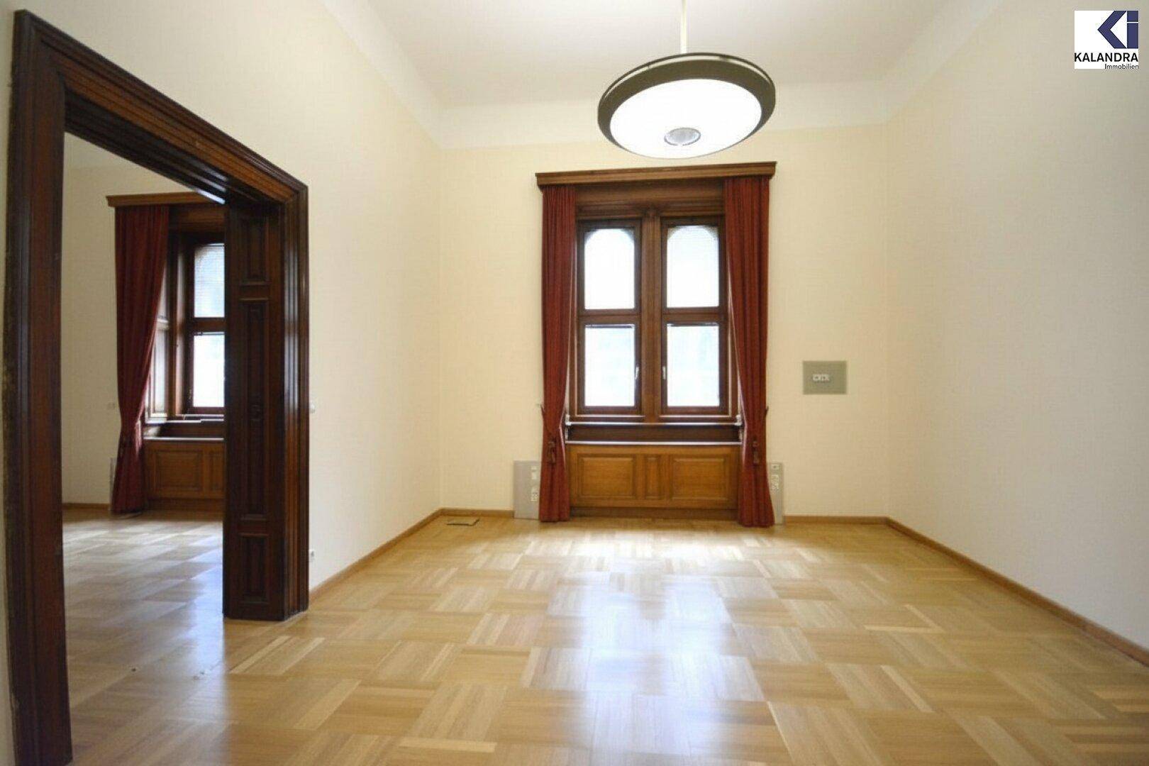 Leerer Raum mit hellem Parkettboden, hohen Decken und einem großen Fenster mit roten Vorhängen.