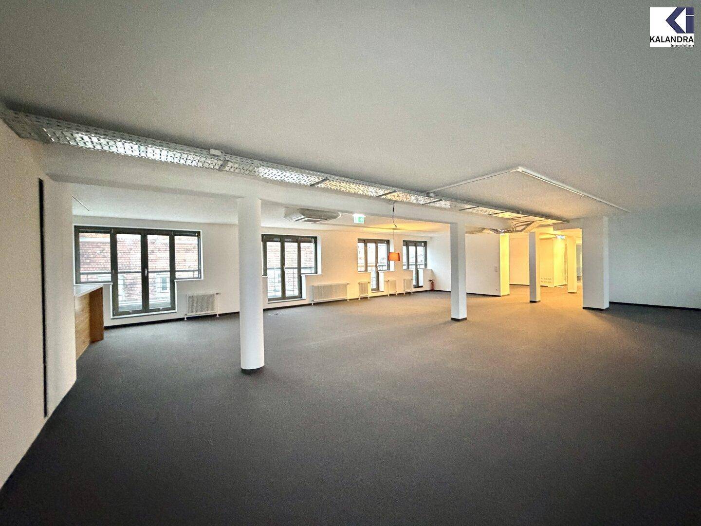 Großer, offener Büroraum mit Teppichboden, Säulen und sichtbaren Deckeninstallationen, hell durch große Fenster.