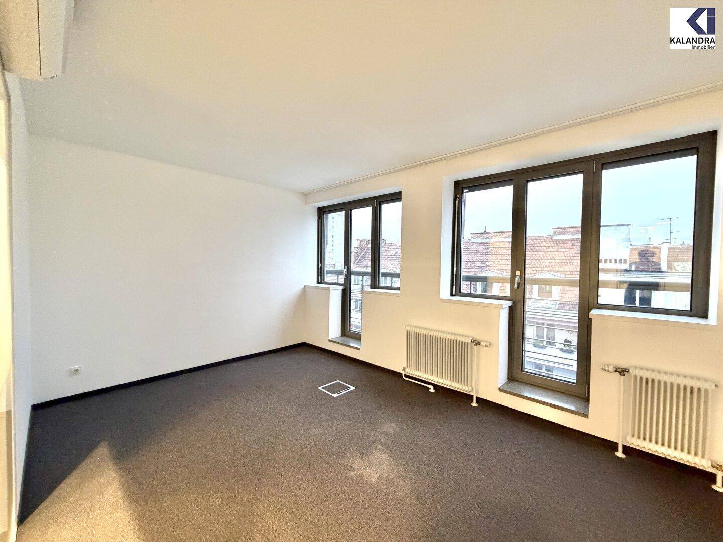 Heller Büroraum mit Teppichboden und zwei großen Fenstertüren, die viel Tageslicht hereinlassen.