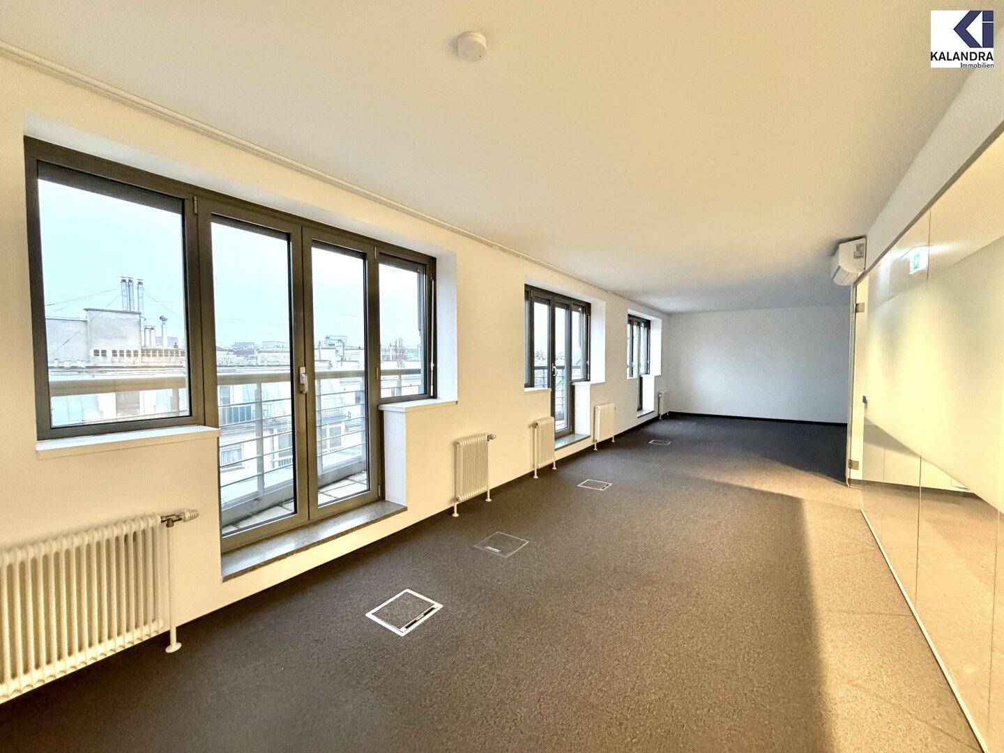 Heller Büroraum mit Teppichboden, großen Fenstern und Heizkörpern, ausgestattet mit Bodentanks für Anschlüsse.