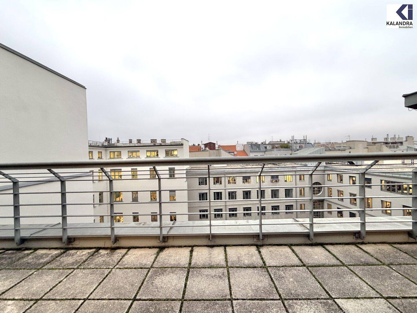 Dachterrasse mit Fliesenboden und Blick auf die umliegenden Stadtgebäude an einem bewölkten Tag.