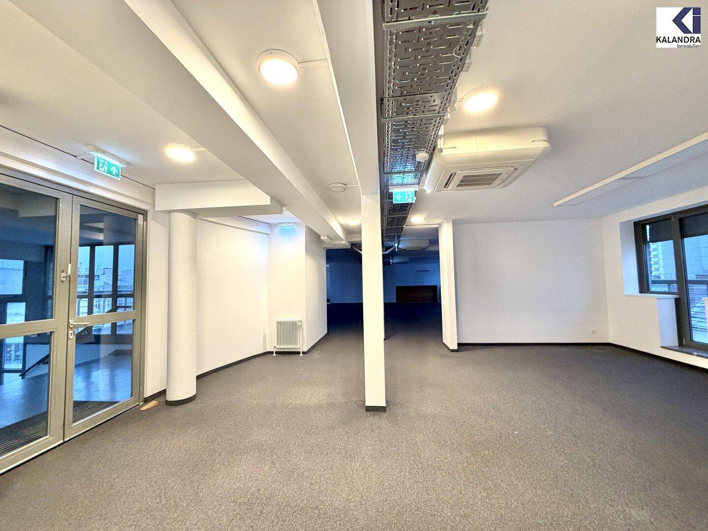 Geräumiger Bürobereich mit Teppichboden, Säulen und sichtbaren Deckeninstallationen, mit Zugang durch eine Glastür.