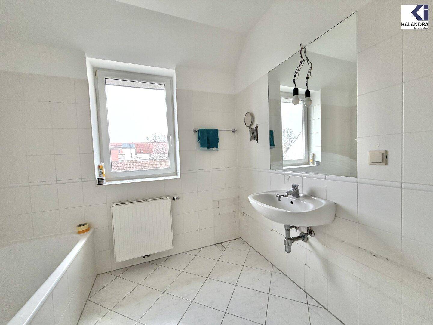 Helles Badezimmer mit Badewanne, Waschbecken, großem Spiegel und Fenster für natürliche Belüftung.