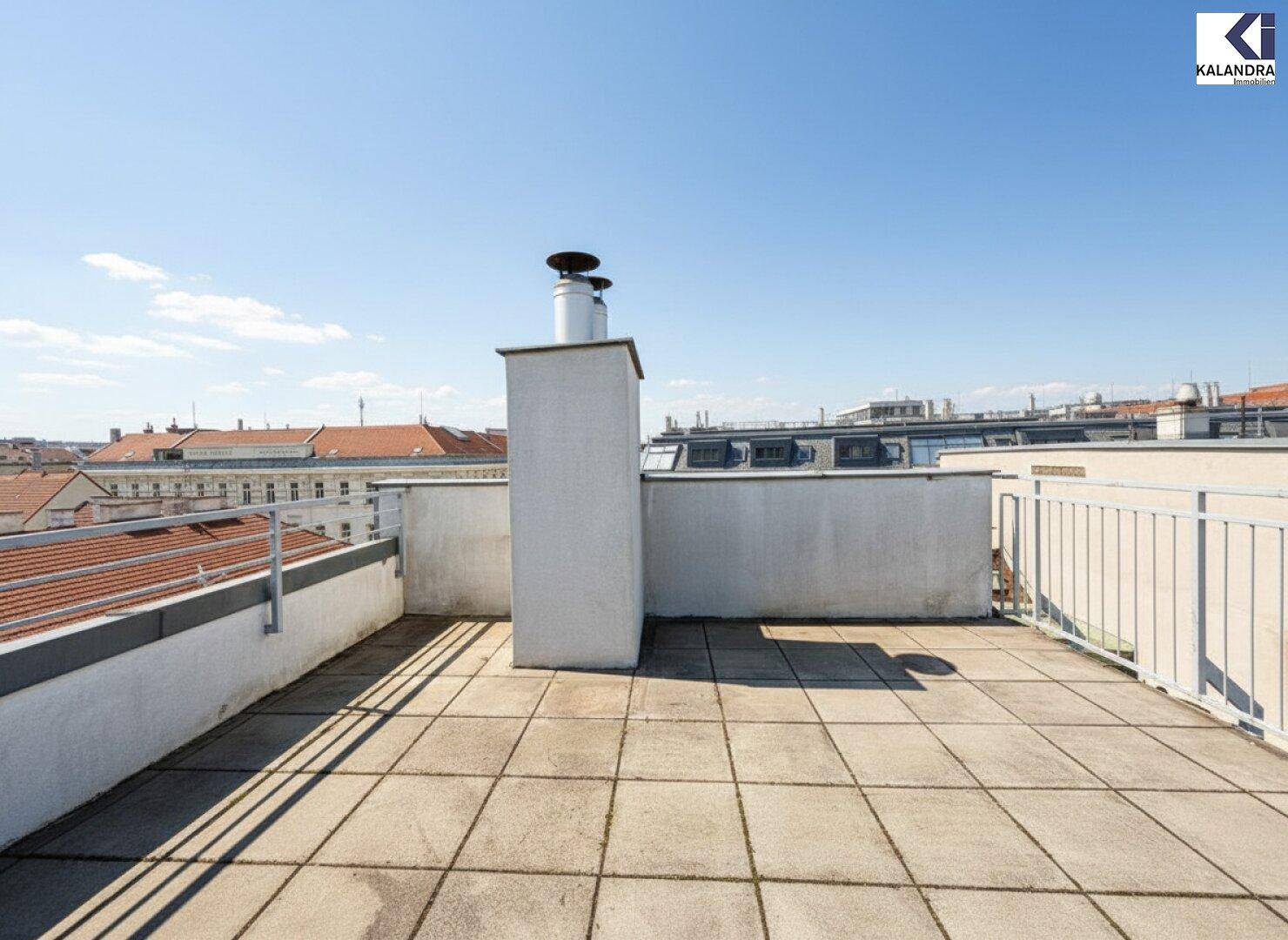 Geräumige Dachterrasse mit Fliesenboden und Blick über die Dächer der Stadt an einem sonnigen Tag.