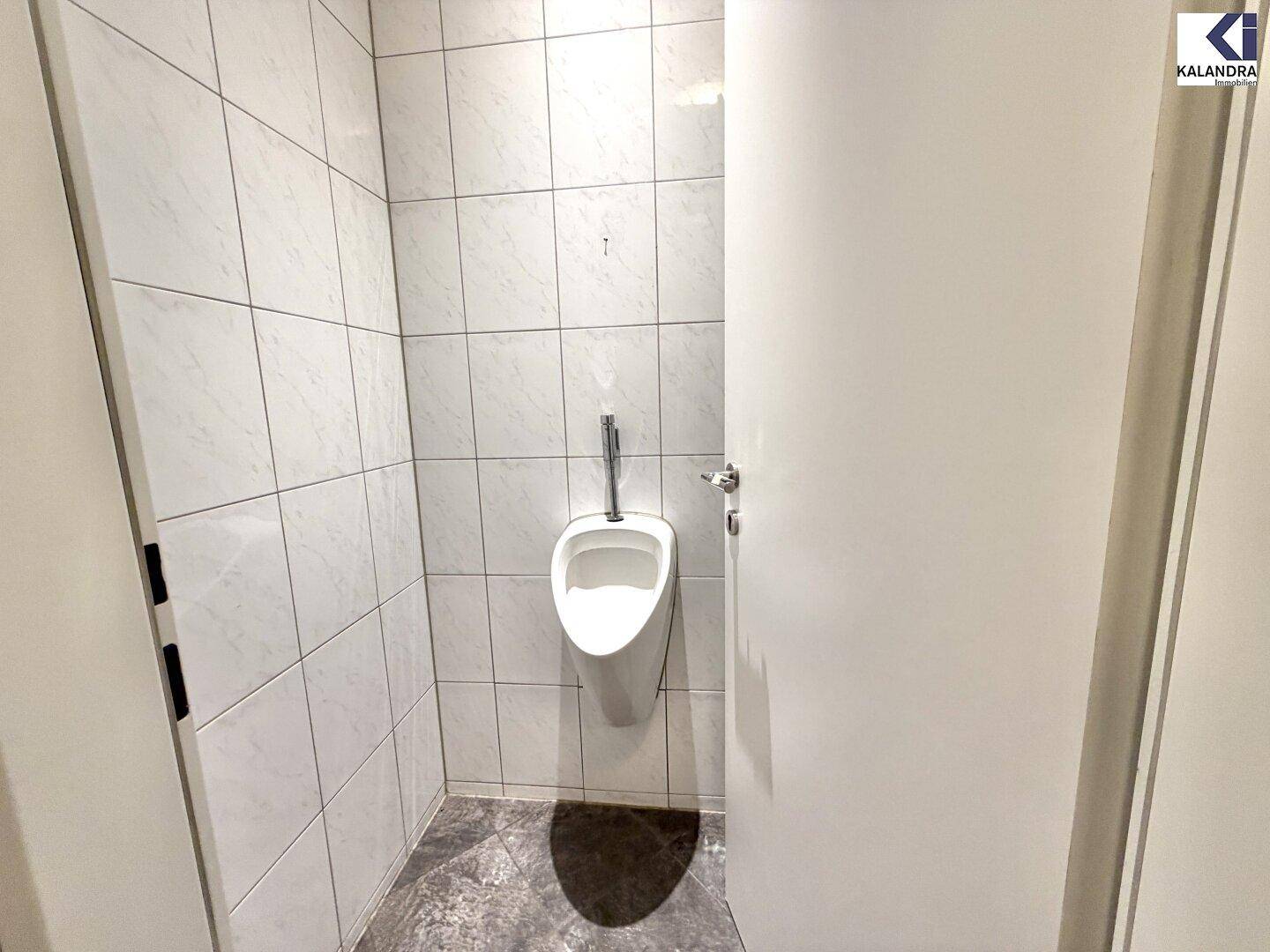 Toilettenraum mit weißen Fliesen, Urinal und dunklem Fliesenboden, sehr gepflegt.