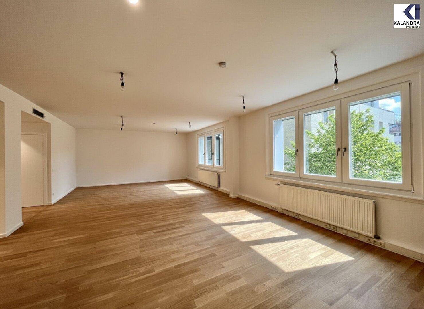 Geräumiger, leerer Raum mit Holzboden, viel natürlichem Licht und mehreren Fenstern mit Blick ins Grüne.