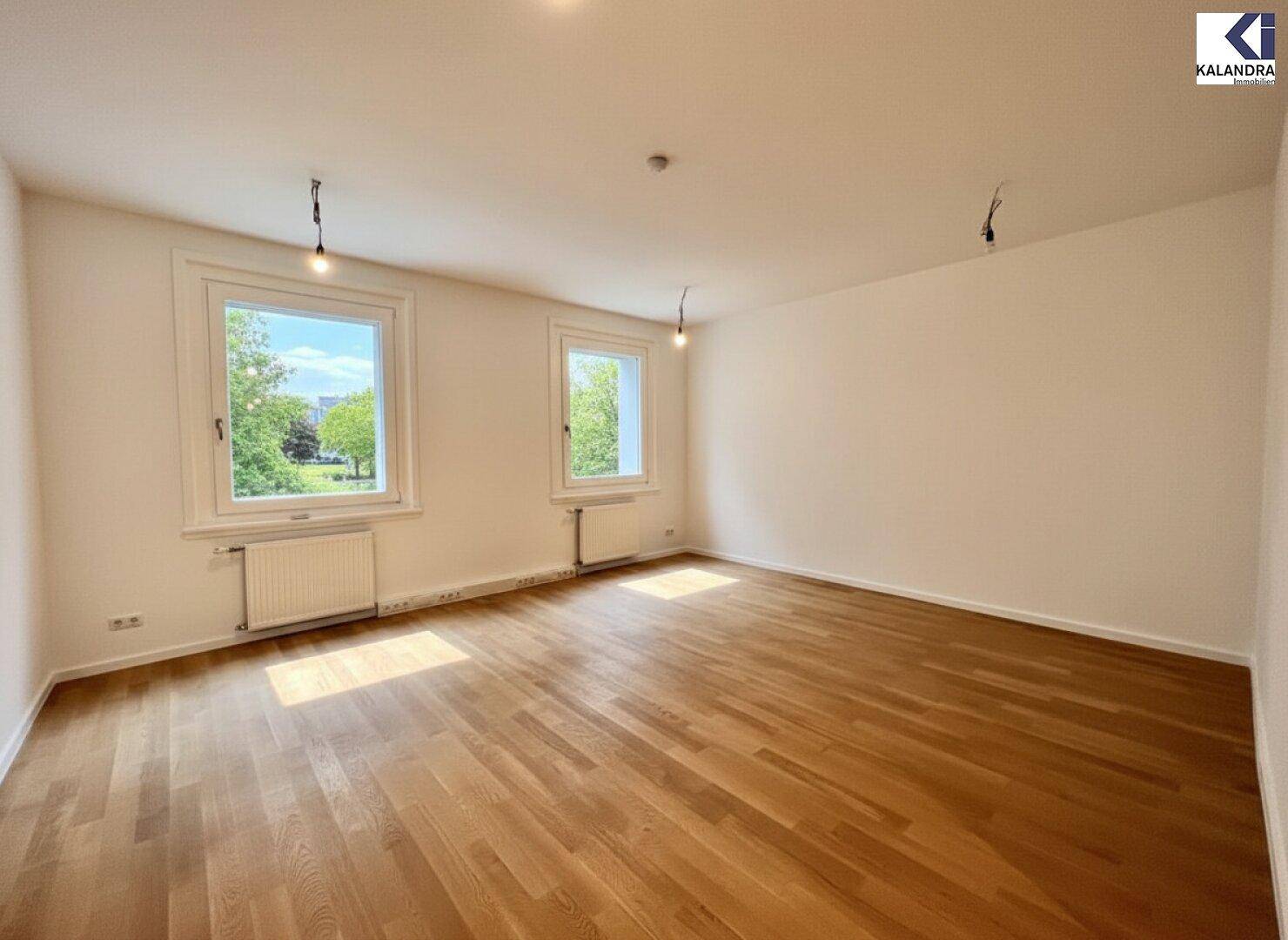 Heller, leerer Raum mit Holzboden und zwei Fenstern, die einen angenehmen Blick ins Grüne bieten.