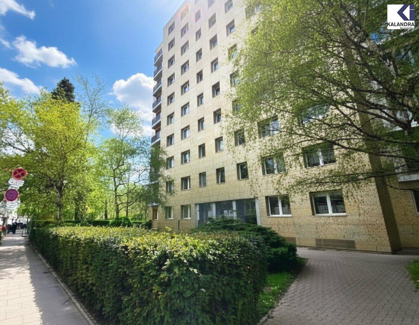 Mehrstöckiges Wohngebäude mit grüner Hecke und Bäumen entlang der Straße unter blauem Himmel.
