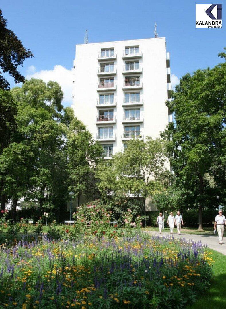 Hohes Wohngebäude mit Balkonen, umgeben von einem blühenden Park mit Bäumen und Spazierwegen.