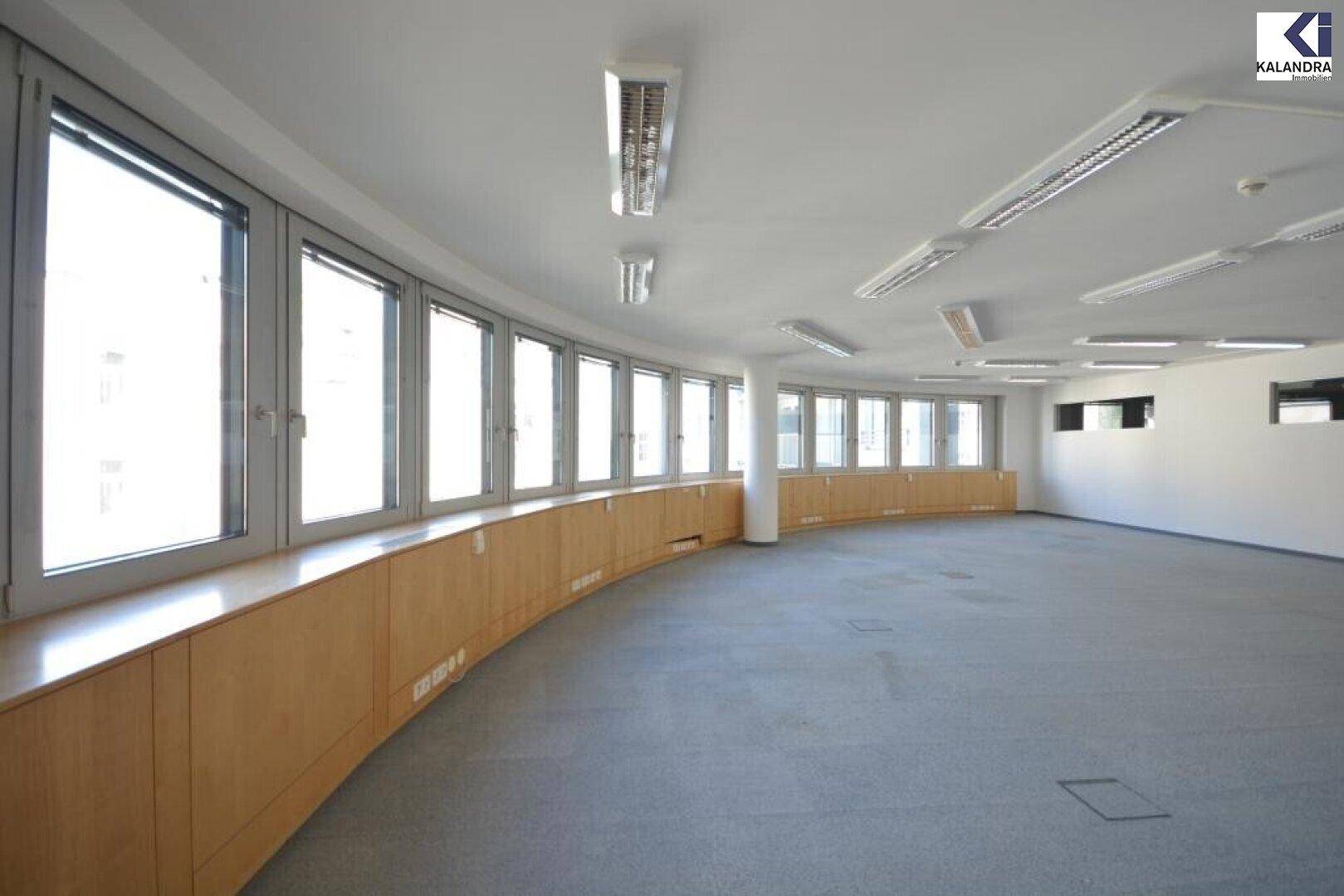 Heller Büroraum mit gebogener Fensterfront, Teppichboden und niedrigen Holzschränken, die viel Tageslicht hereinlassen.