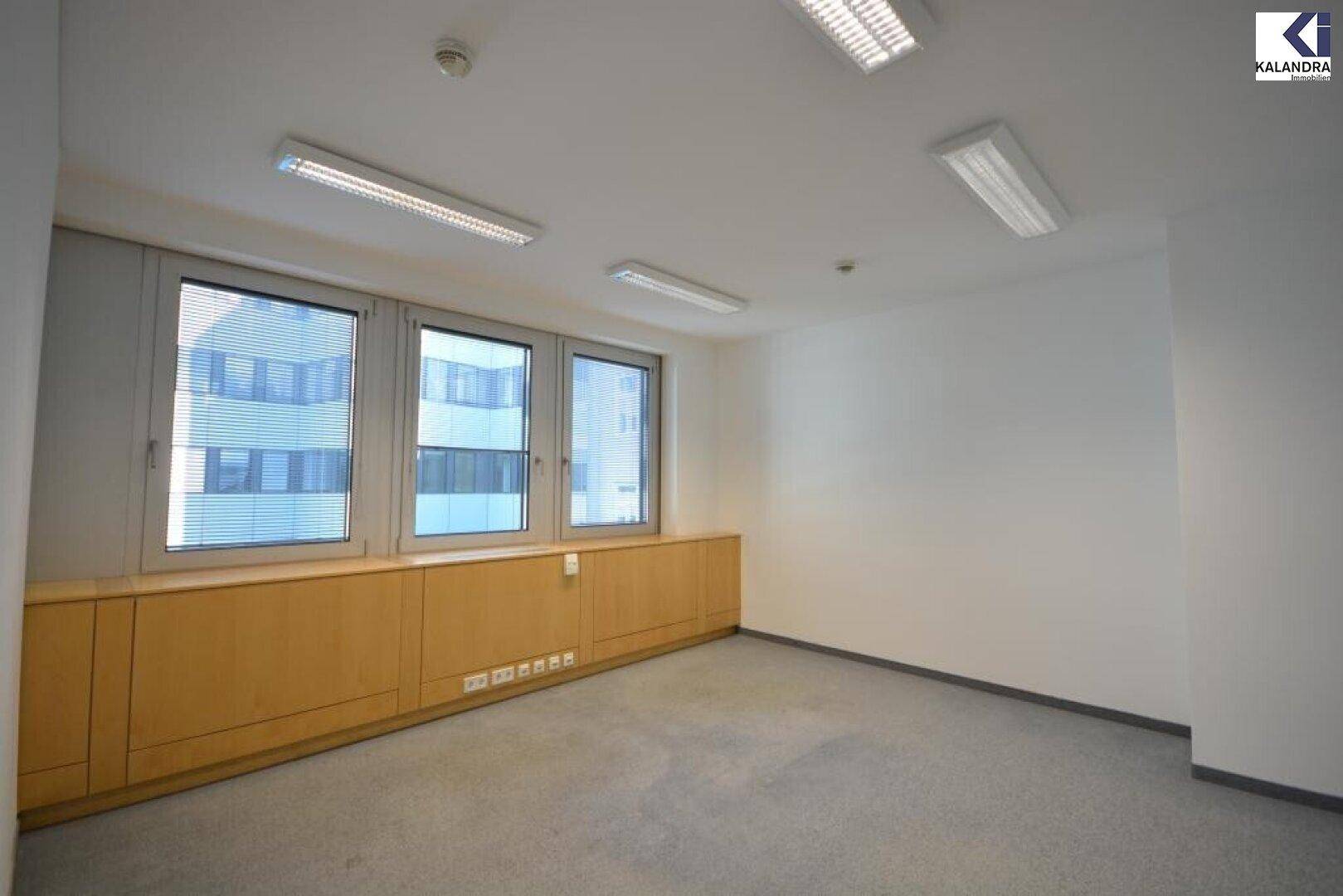Kleinerer Büroraum mit Teppichboden, weißen Wänden und Fenstern mit Jalousien, die Tageslicht hereinlassen.