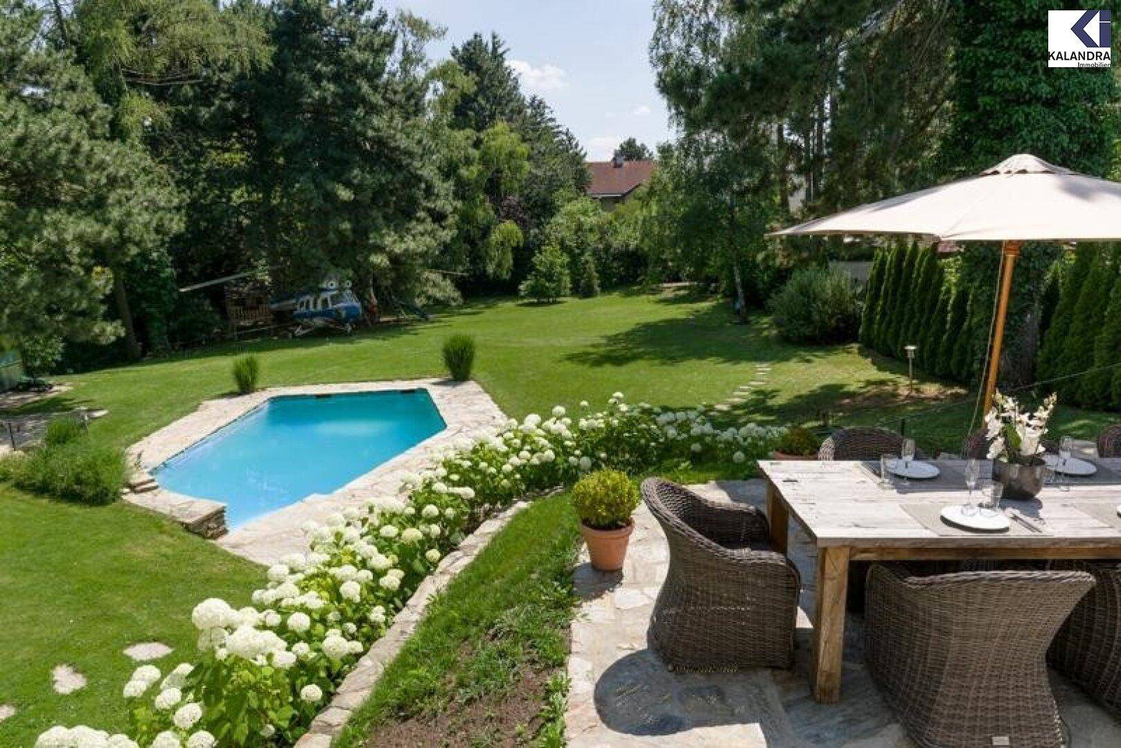 Blick von oben auf den Garten mit Essbereich, Pool und blühenden Sträuchern in sonniger Lage.