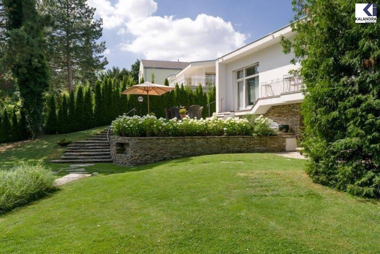Modernes Haus mit großem Garten, Terrasse und gepflegten Hecken unter blauem Himmel.