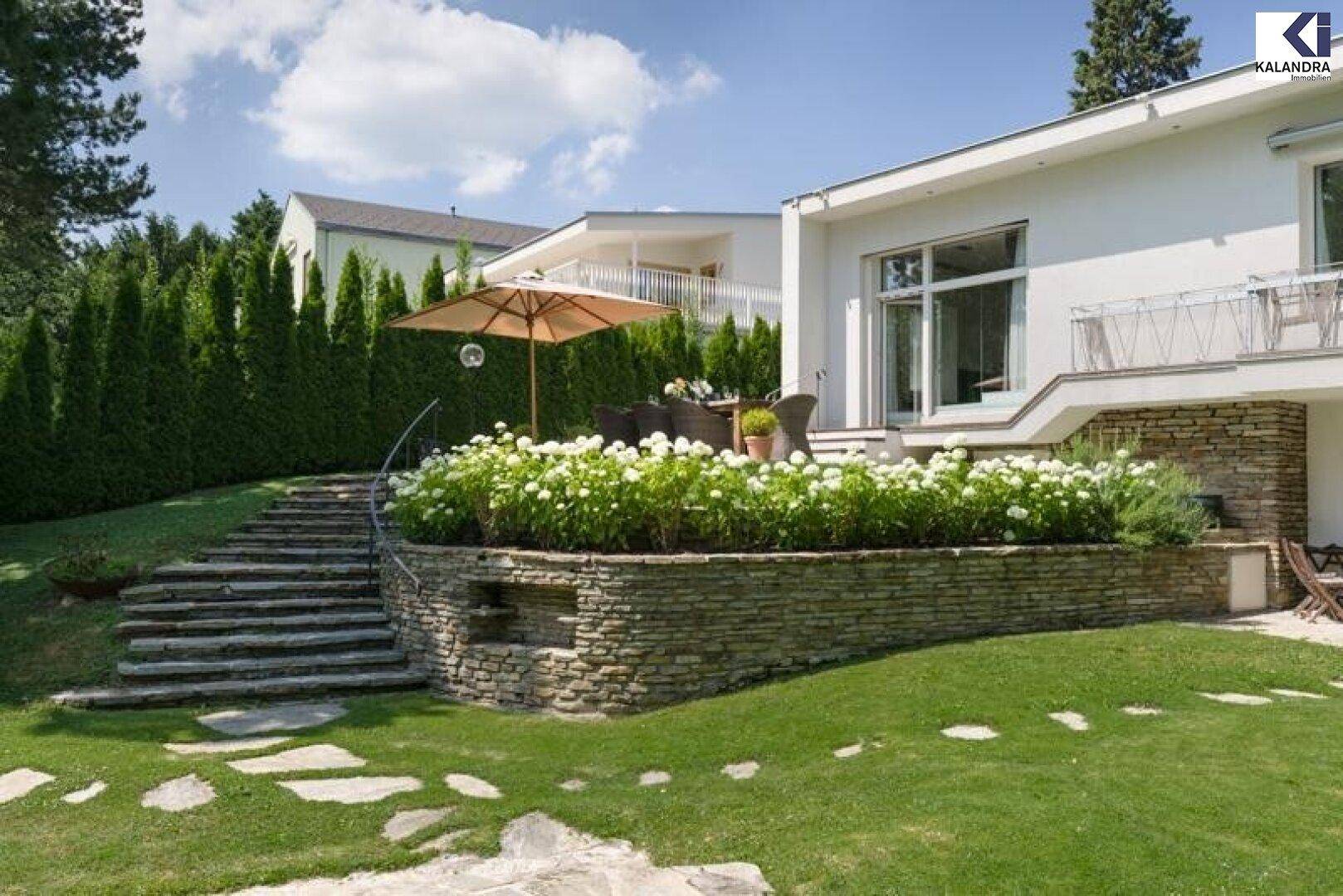 Modernes Haus mit Terrasse, Steintreppe und blühenden Sträuchern im Garten.