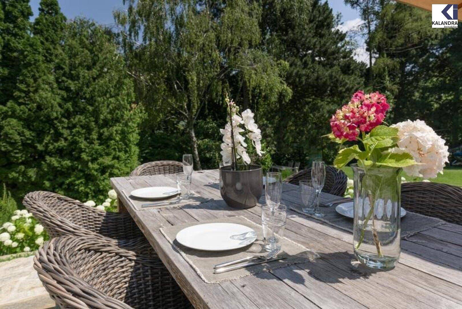 Gedeckter Esstisch auf der Terrasse mit Blumenarrangements und Blick ins Grüne.