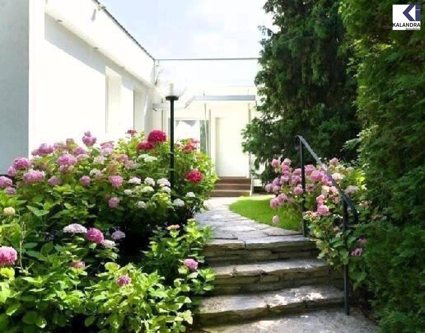 Steinweg zum Eingang, umgeben von blühenden Hortensien und Bäumen.