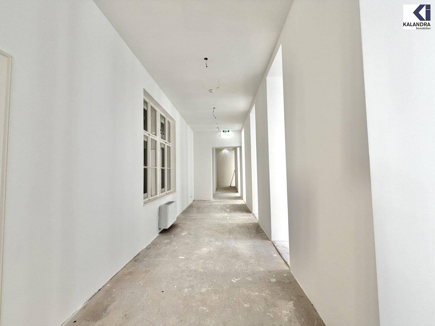 Heller Flur mit einem großen Fenster und unfertigem Betonboden, der zu weiteren Räumen führt.