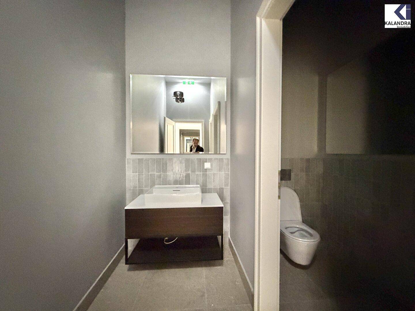 Modernes Badezimmer mit Waschtisch, Spiegel und Toilette, in grauen Farbtönen und Fliesen gehalten.
