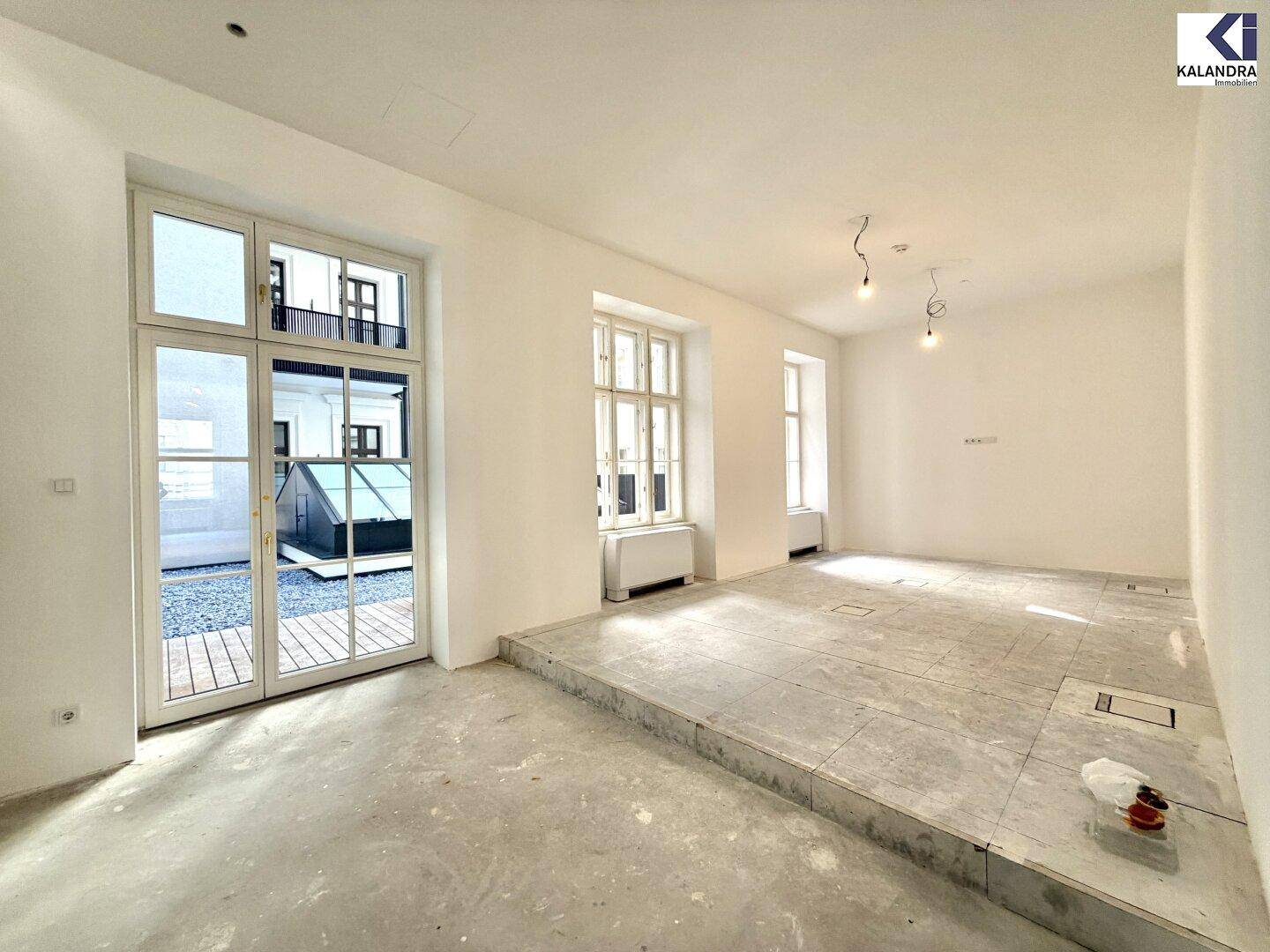 Heller Raum mit großen Fenstertüren, die zu einem Außenbereich führen, und unfertigem Boden.