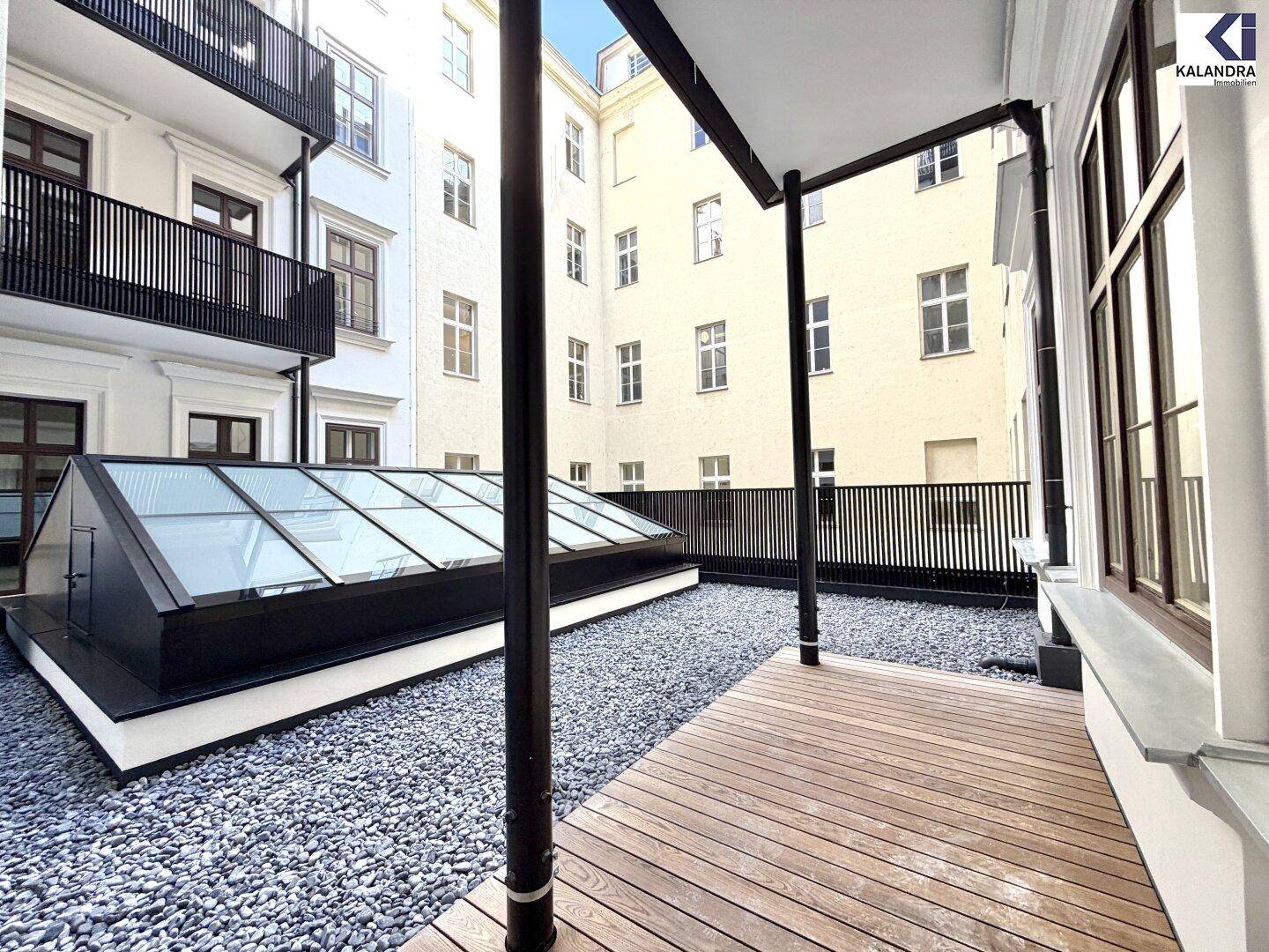 Attraktive Terrasse mit Holzboden, Kiesfläche und Glasdach, umgeben von historischen Gebäuden im Innenhof.