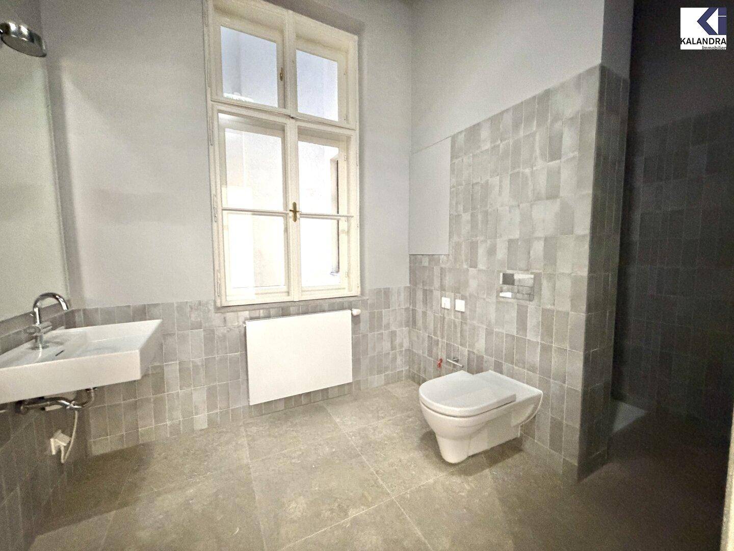 Helles Badezimmer mit Fenster, Waschtisch und wandhängender Toilette, in grauen Fliesen gehalten.