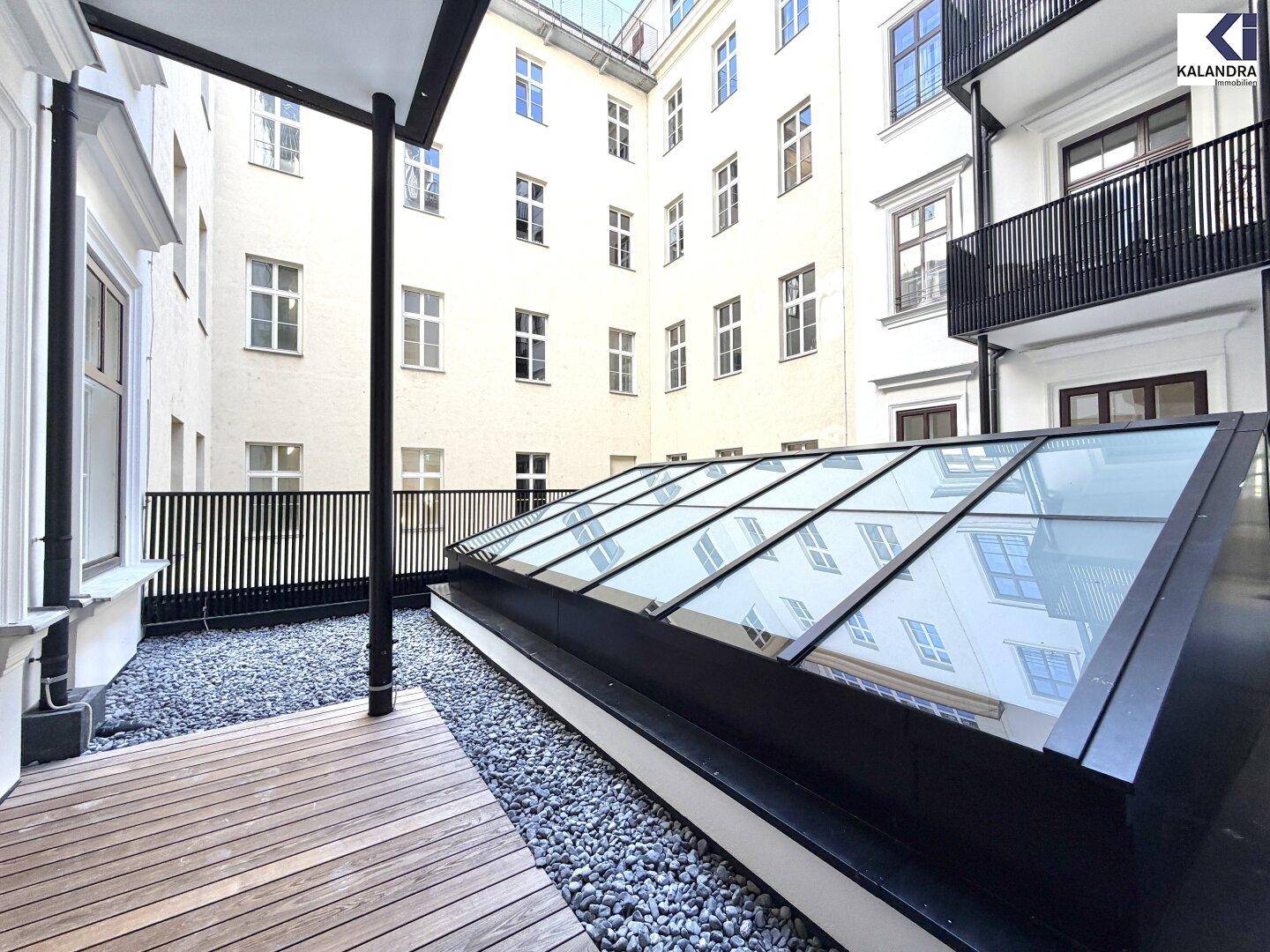 Detailansicht des Innenhofs mit Holzterrasse, Kies und einem modernen Glasdach, umgeben von Gebäuden.