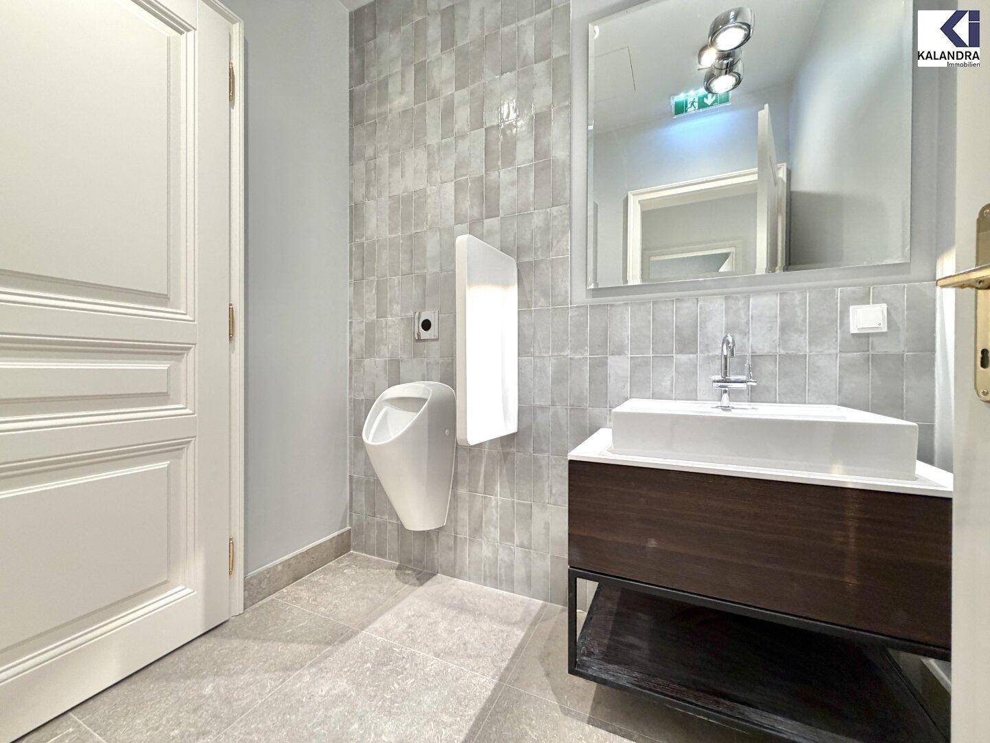 Modernes Gäste-WC mit Urinal, Waschbecken und großem Spiegel, gefliest in Grau.