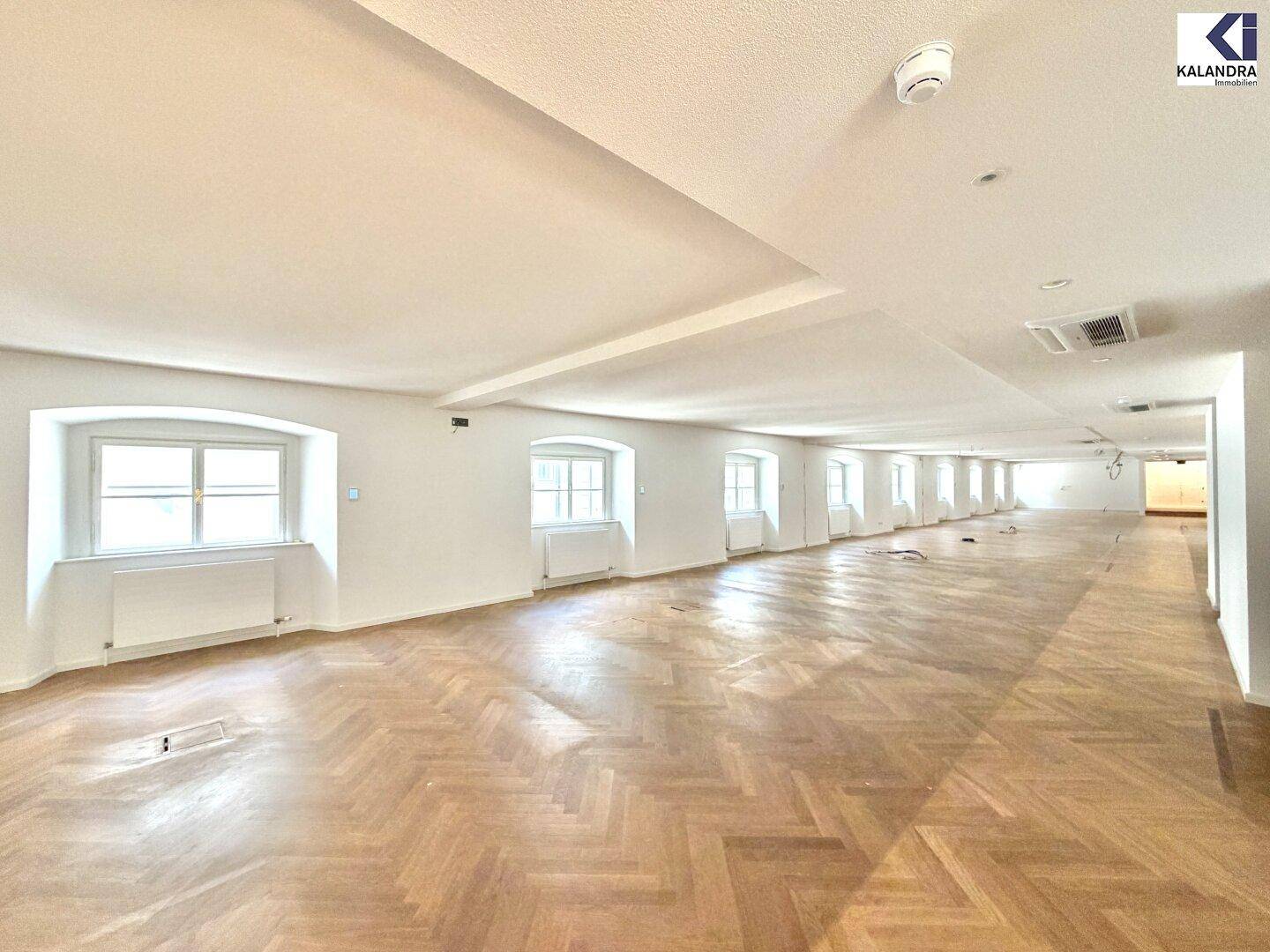 Langer, heller Raum mit Fischgrätparkett und einer Reihe von Fenstern, die für viel Tageslicht sorgen.