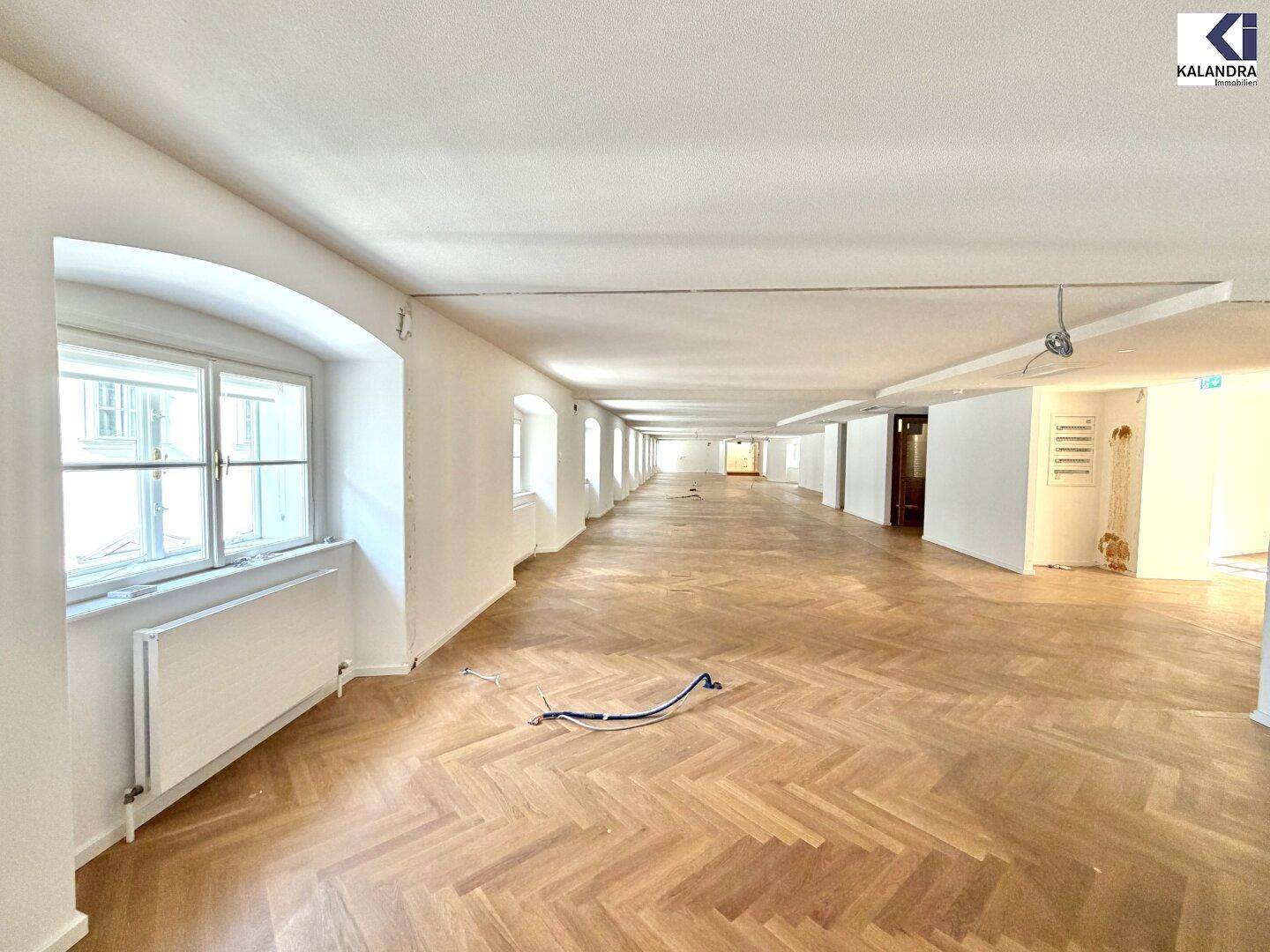 Geräumiger, heller Büroraum mit Fischgrätparkett und einer langen Fensterfront, die den Raum erhellt.