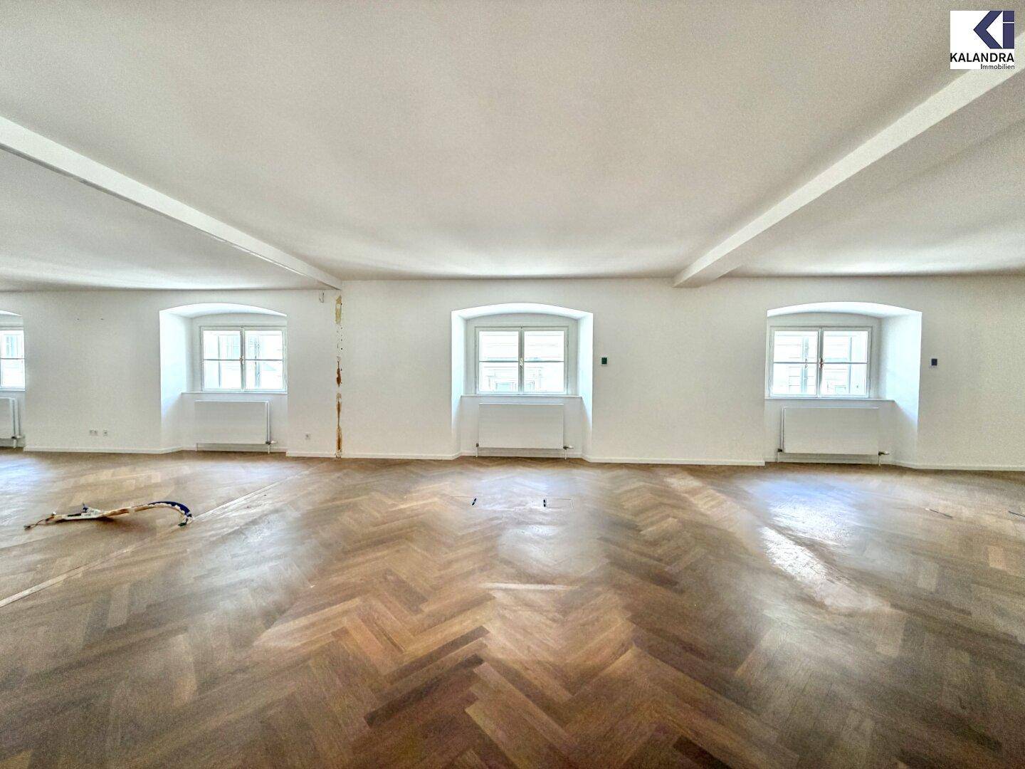 Weitläufiger, heller Raum mit Fischgrätparkett und drei Fenstern, die den Raum mit Tageslicht durchfluten.