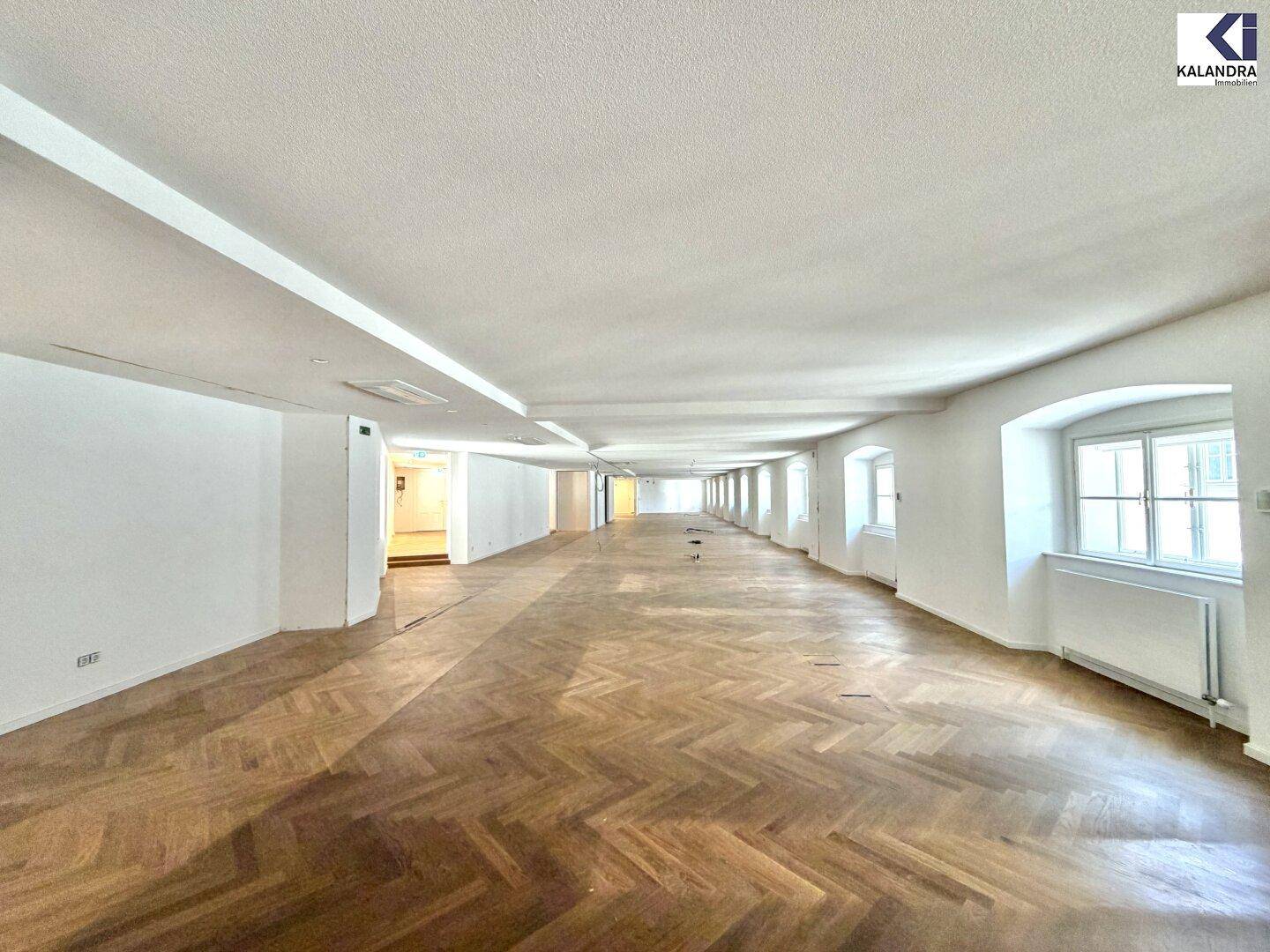 Langer, heller Raum mit Fischgrätparkett und einer Reihe von Fenstern, die für viel Tageslicht sorgen.