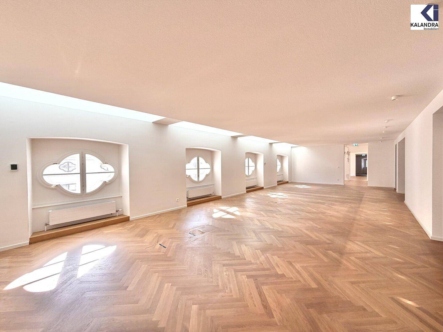 Langer, heller Raum mit Fischgrätparkett und einer Reihe von Fenstern, die für viel Tageslicht sorgen.