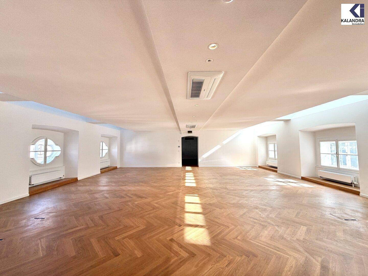 Großer, leerer Raum mit elegantem Fischgrätparkett und mehreren Fenstern, darunter ein markantes Rundfenster.