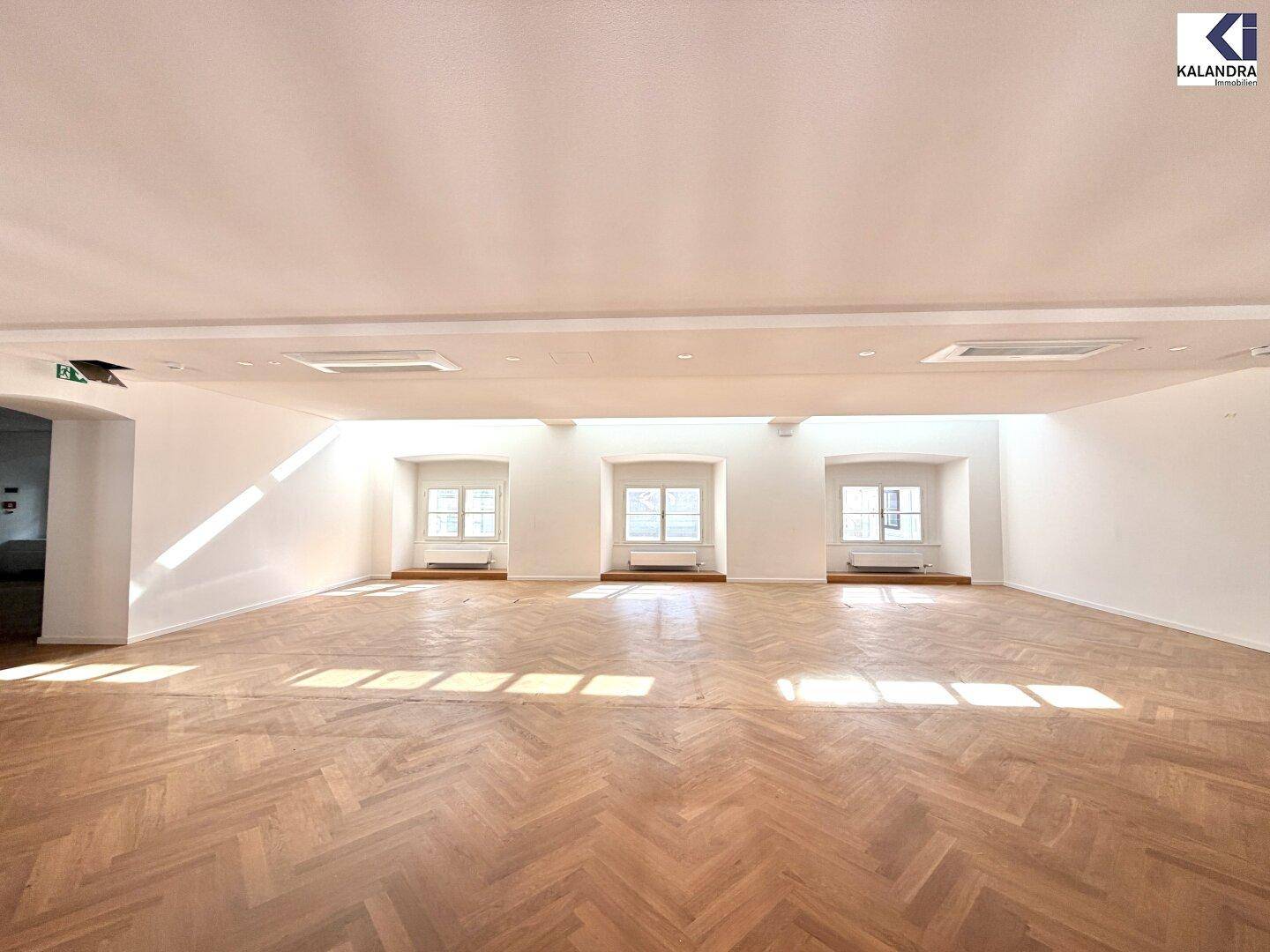 Heller, weitläufiger Raum mit klassischem Fischgrätparkett und drei großen Fenstern, die viel Tageslicht hereinlassen.
