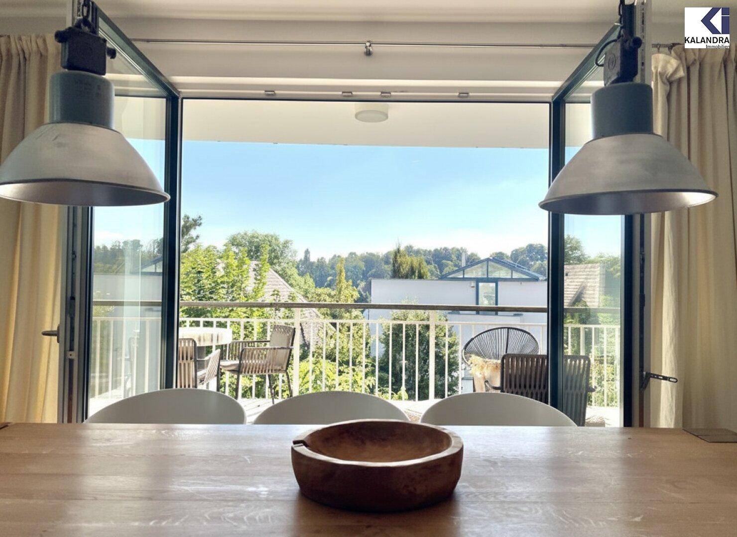 Essbereich mit Holztisch und Blick durch große Fenster auf den sonnigen Balkon und die grüne Umgebung.