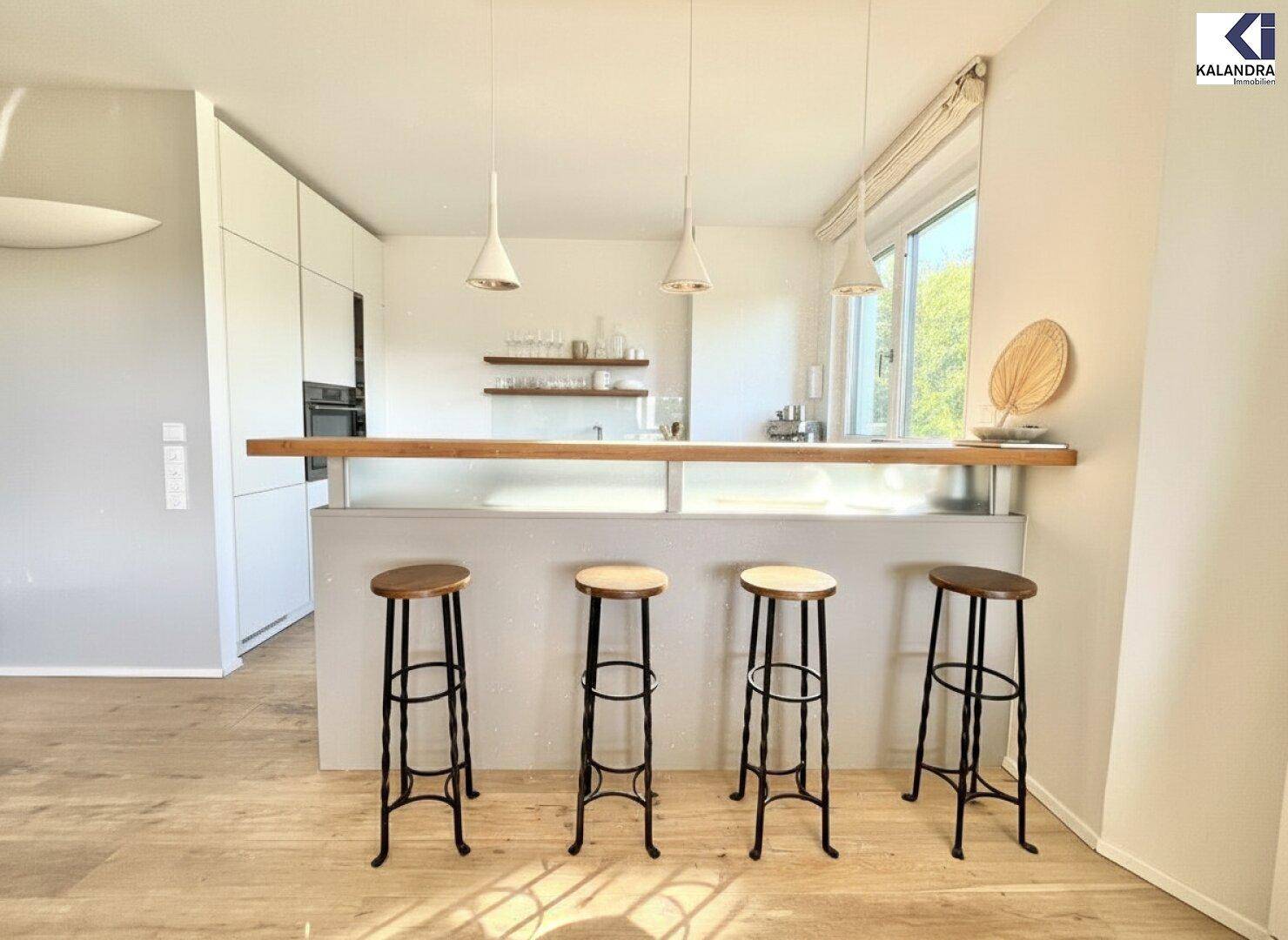 Moderne Küche mit weißer Front, Holztheke und Barhockern, offen zum Wohnbereich.