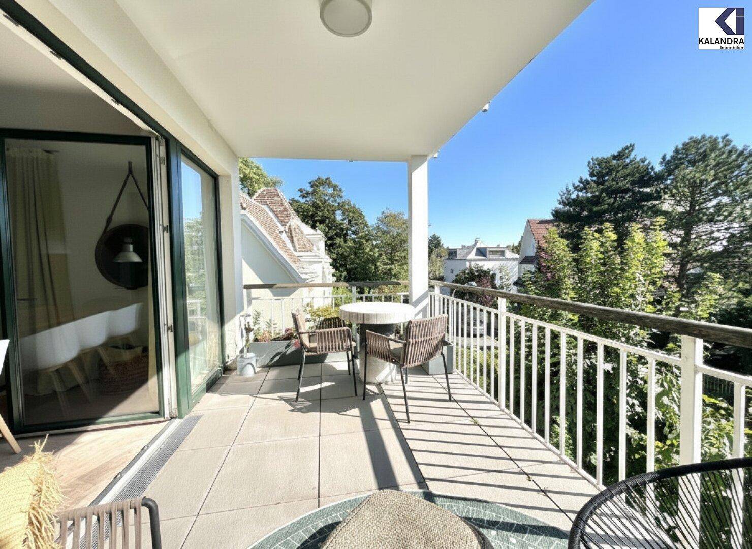 Heller Balkon mit Fliesenboden und Blick auf die grünen Bäume und Nachbarhäuser.