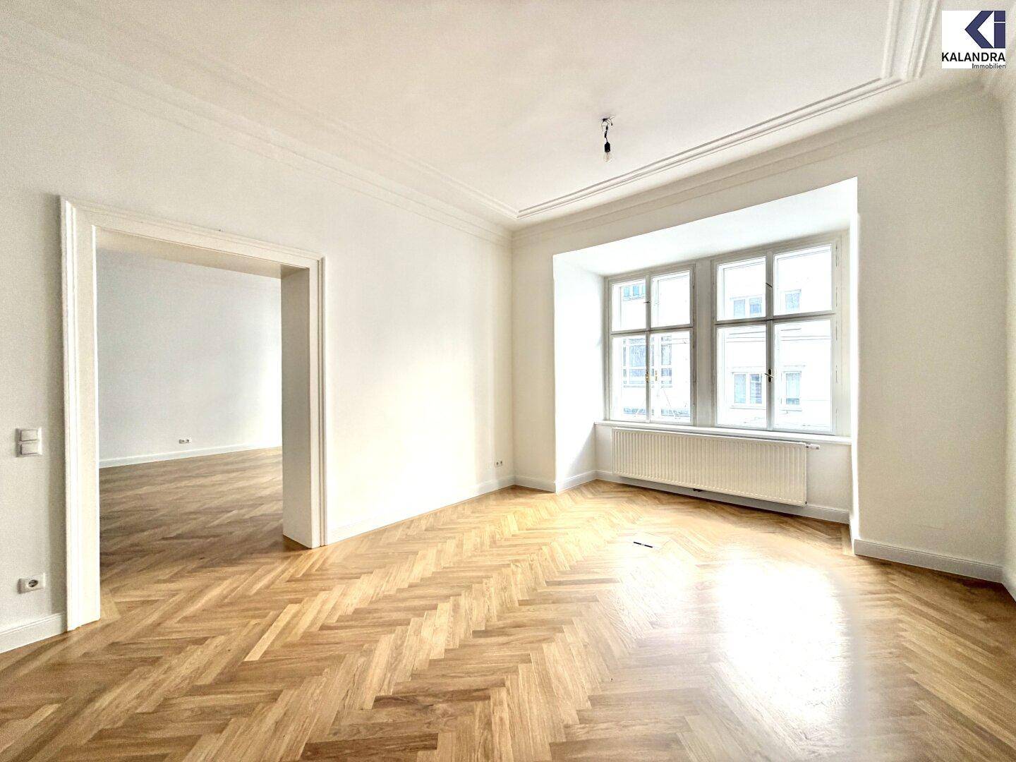 Heller Wohnraum mit klassischem Fischgrätparkett, hohen Decken und einem großen Fenster, das viel Tageslicht hereinlässt.