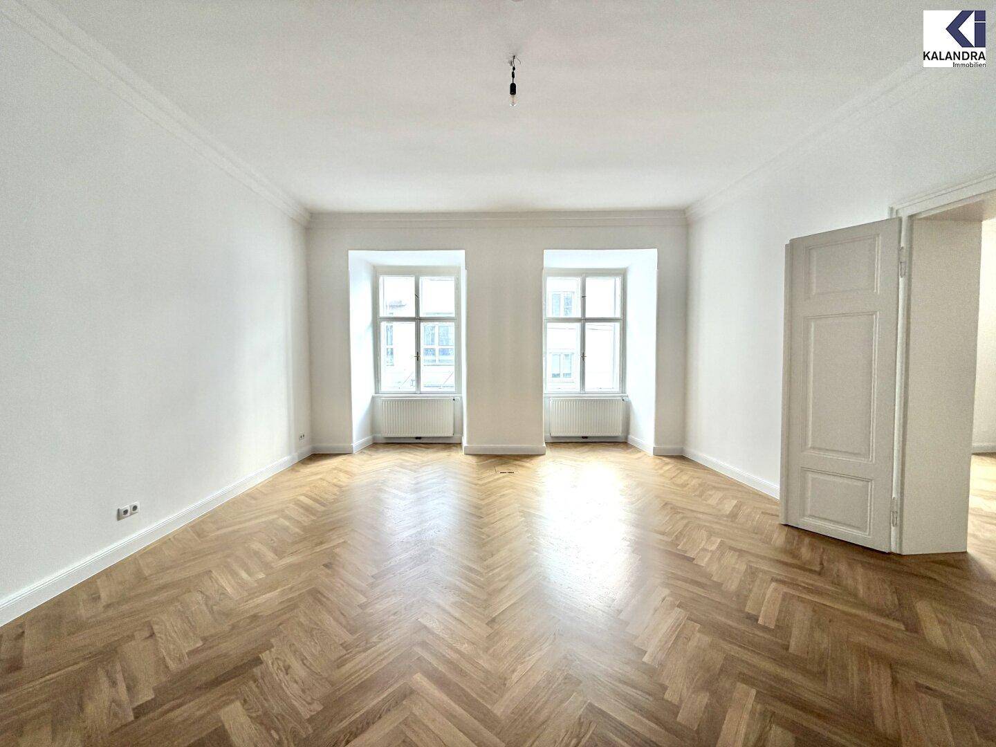 Geräumiges Zimmer mit zwei großen Fenstern, klassischem Fischgrätparkett und hohen Decken, ideal für Wohn- oder Arbeitszwecke.