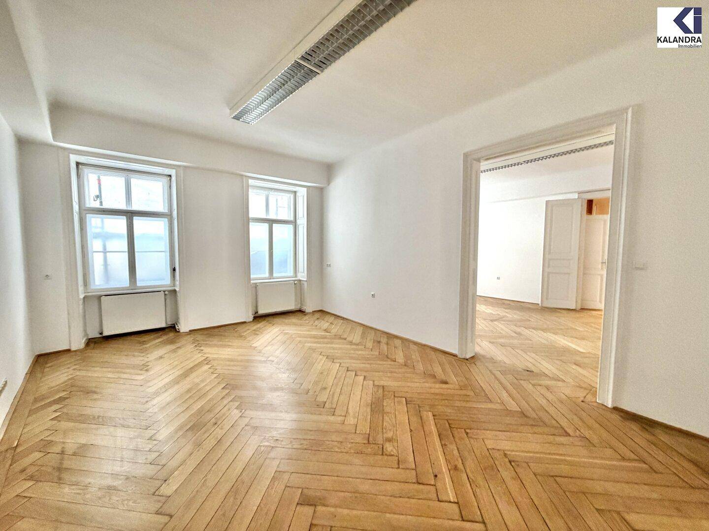 Großes Zimmer mit Fischgrätparkett und Zugang zu einem weiteren Raum, hell durch zwei hohe Fenster.