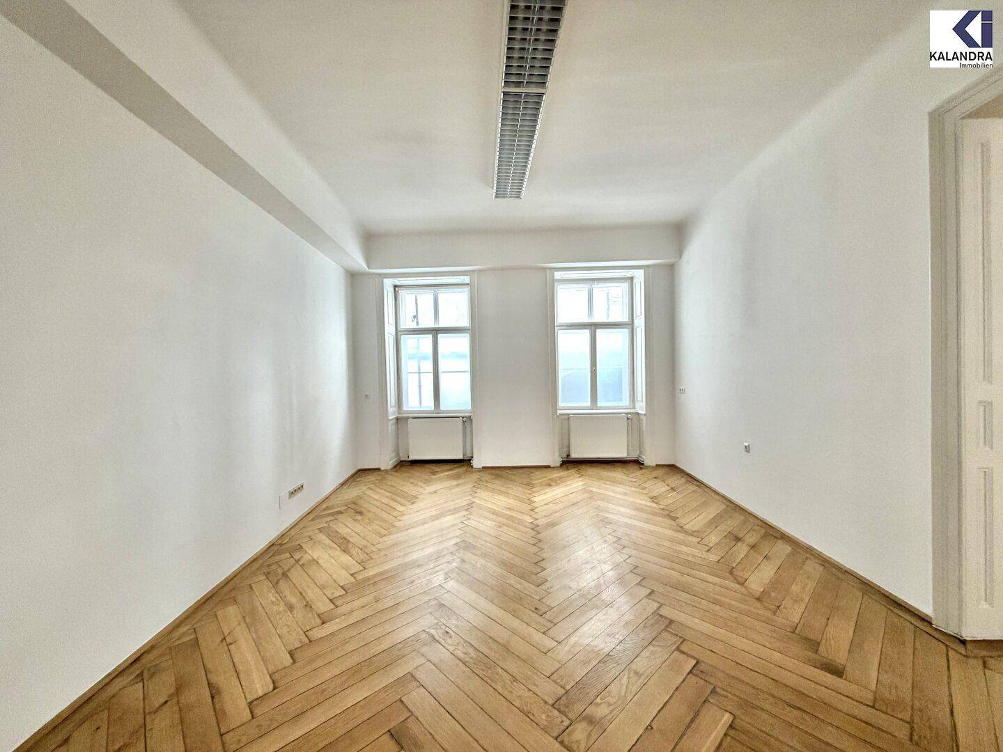 Leeres, helles Zimmer mit elegantem Fischgrätparkett und zwei hohen Fenstern, ideal für vielfältige Nutzung.