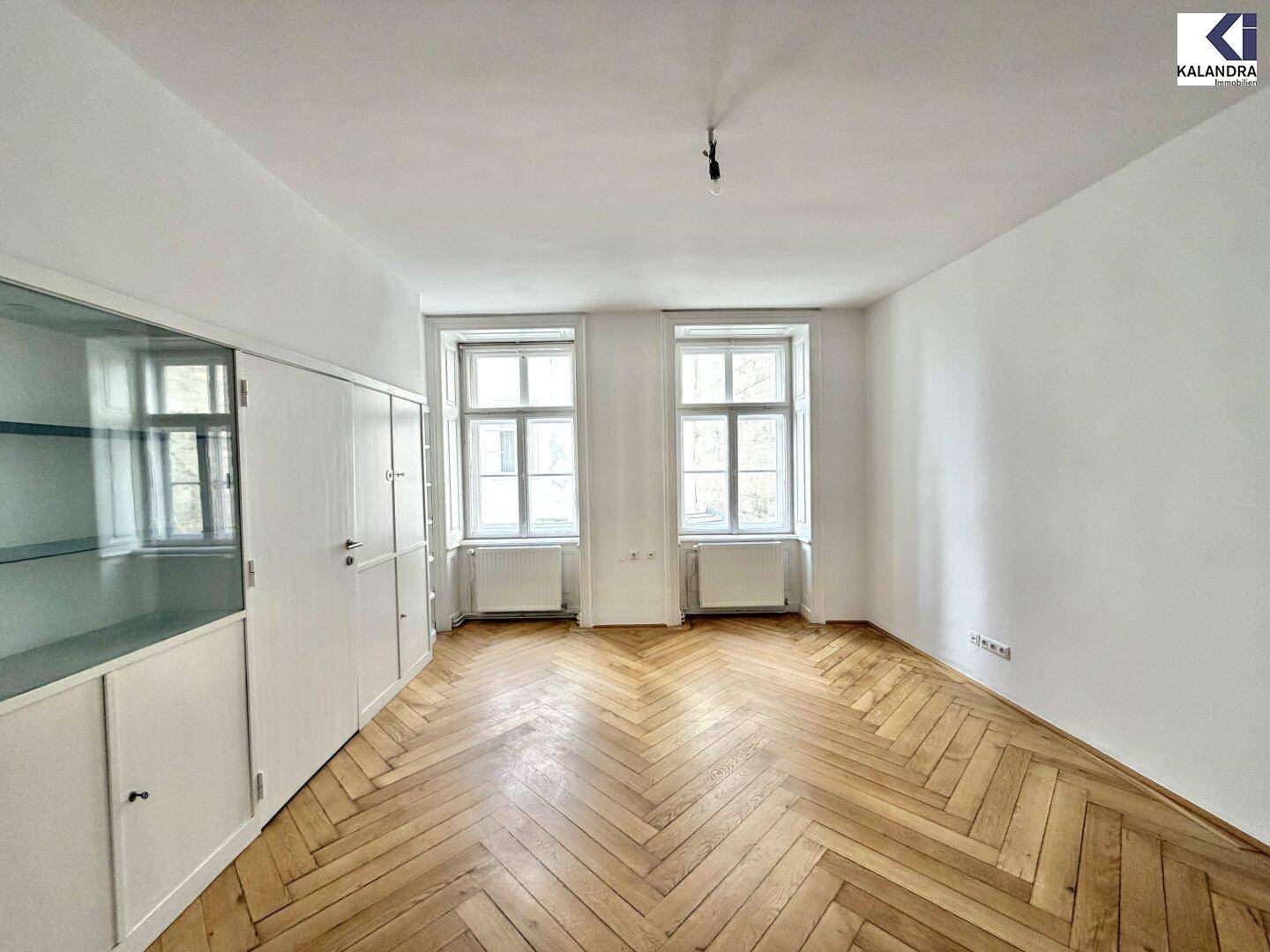 Geräumiges Zimmer mit Fischgrätparkett, zwei großen Fenstern und Einbauschränken, ideal als Wohn- oder Arbeitsbereich.