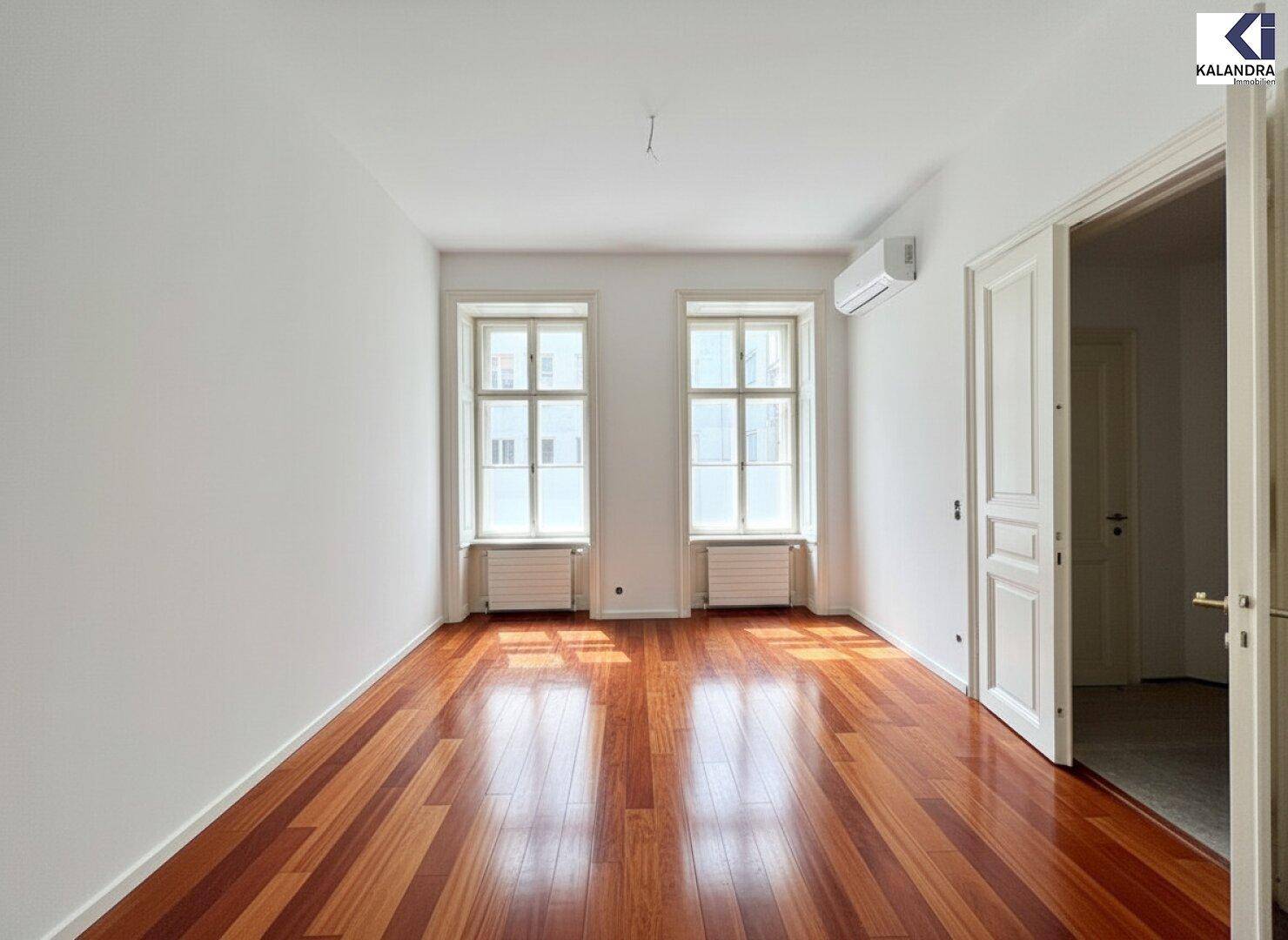 Heller Raum mit Holzboden, Klimaanlage und einer geöffneten Tür, die den Blick in einen weiteren Bereich freigibt.