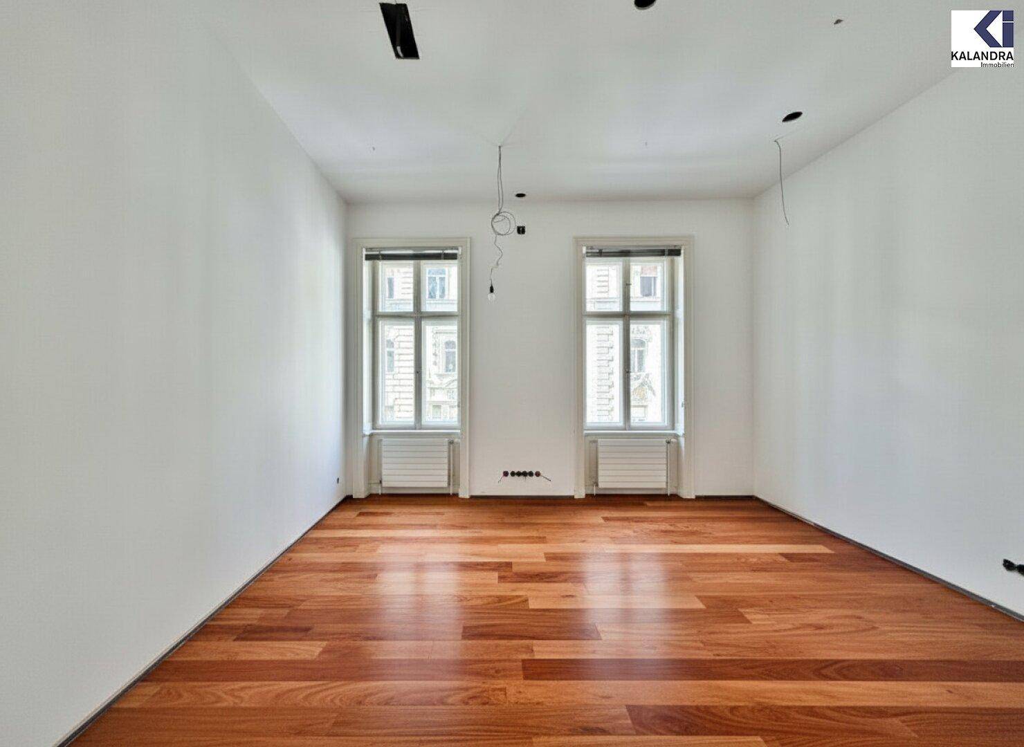 Unmöbliertes Zimmer mit glänzendem Holzfußboden und zwei Fenstern, die den Raum mit natürlichem Licht füllen.