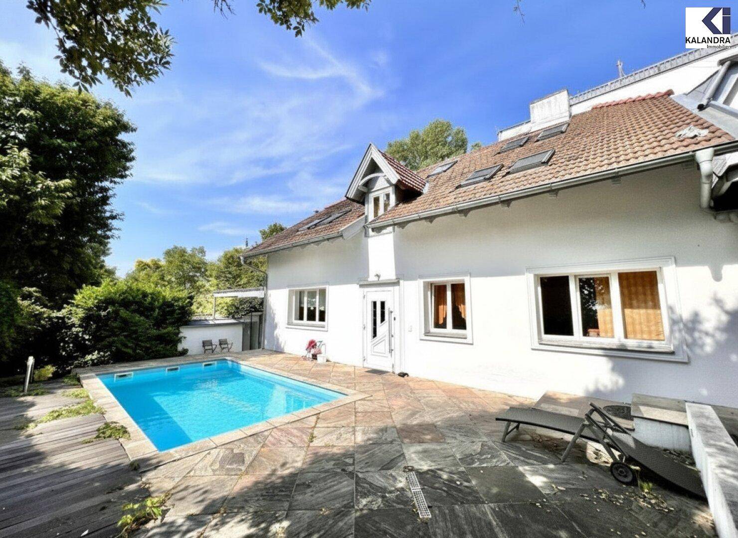 Weitläufiger Poolbereich mit angrenzender Terrasse und der weißen Fassade des Hauses im Sonnenlicht.