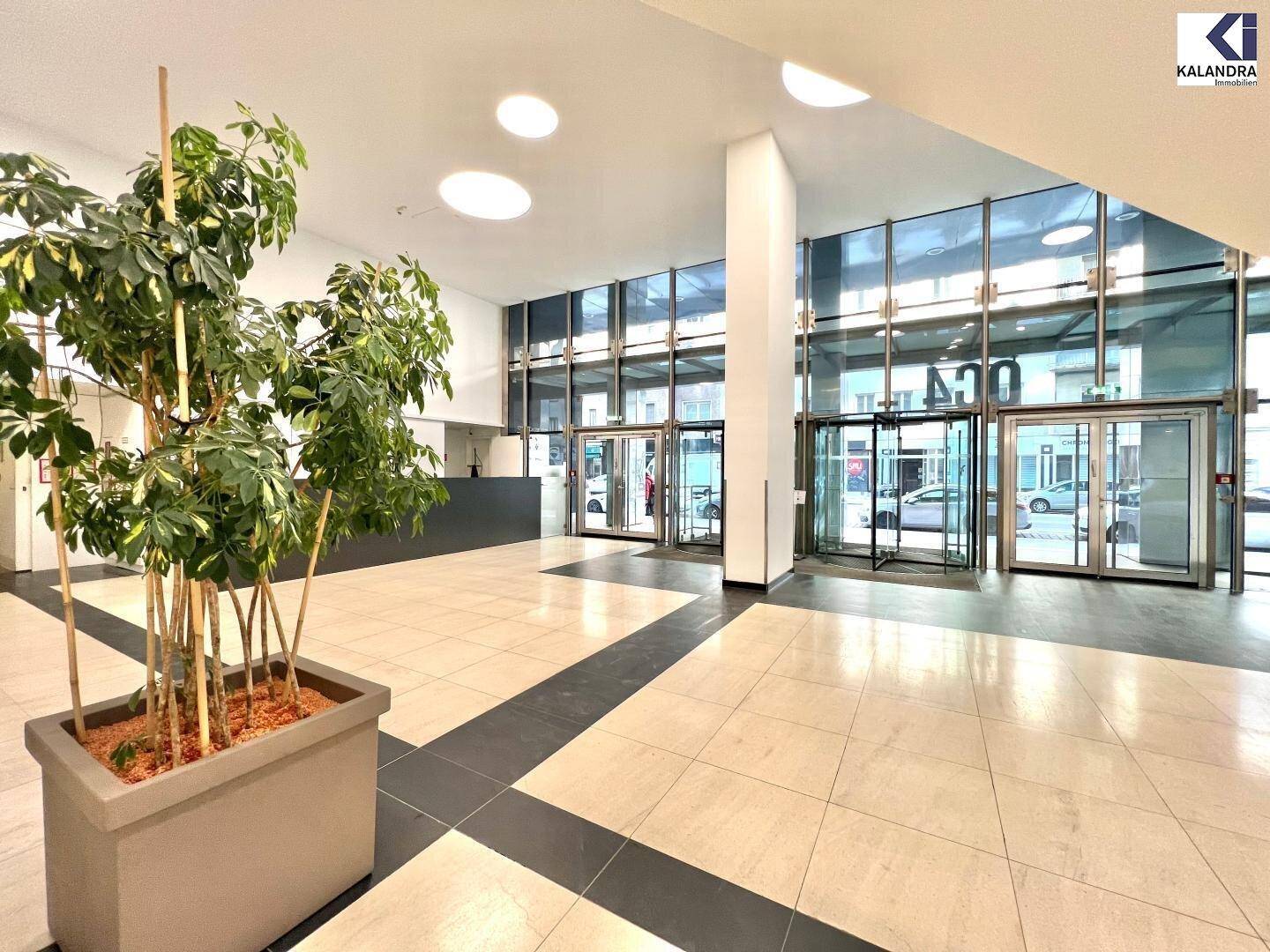 Geräumige und helle Lobby mit großen Glasfronten, Fliesenboden und einer dekorativen Pflanze im Eingangsbereich.